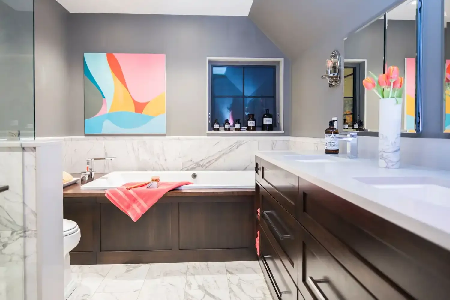 Modern bathroom with gray walls, a white marble countertop, dark wooden cabinets, a white marble bathtub with a pink towel, and a colorful abstract painting on the wall.