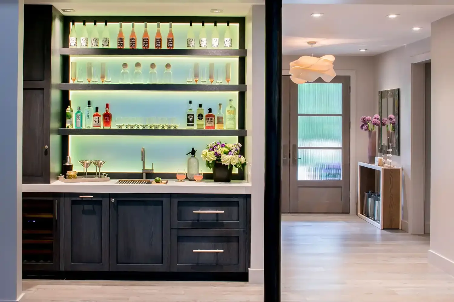 Modern home bar area with black cabinetry, illuminated shelves displaying bottles of alcohol, glasses, and flowers, with a hallway and door with frosted glass in the background.