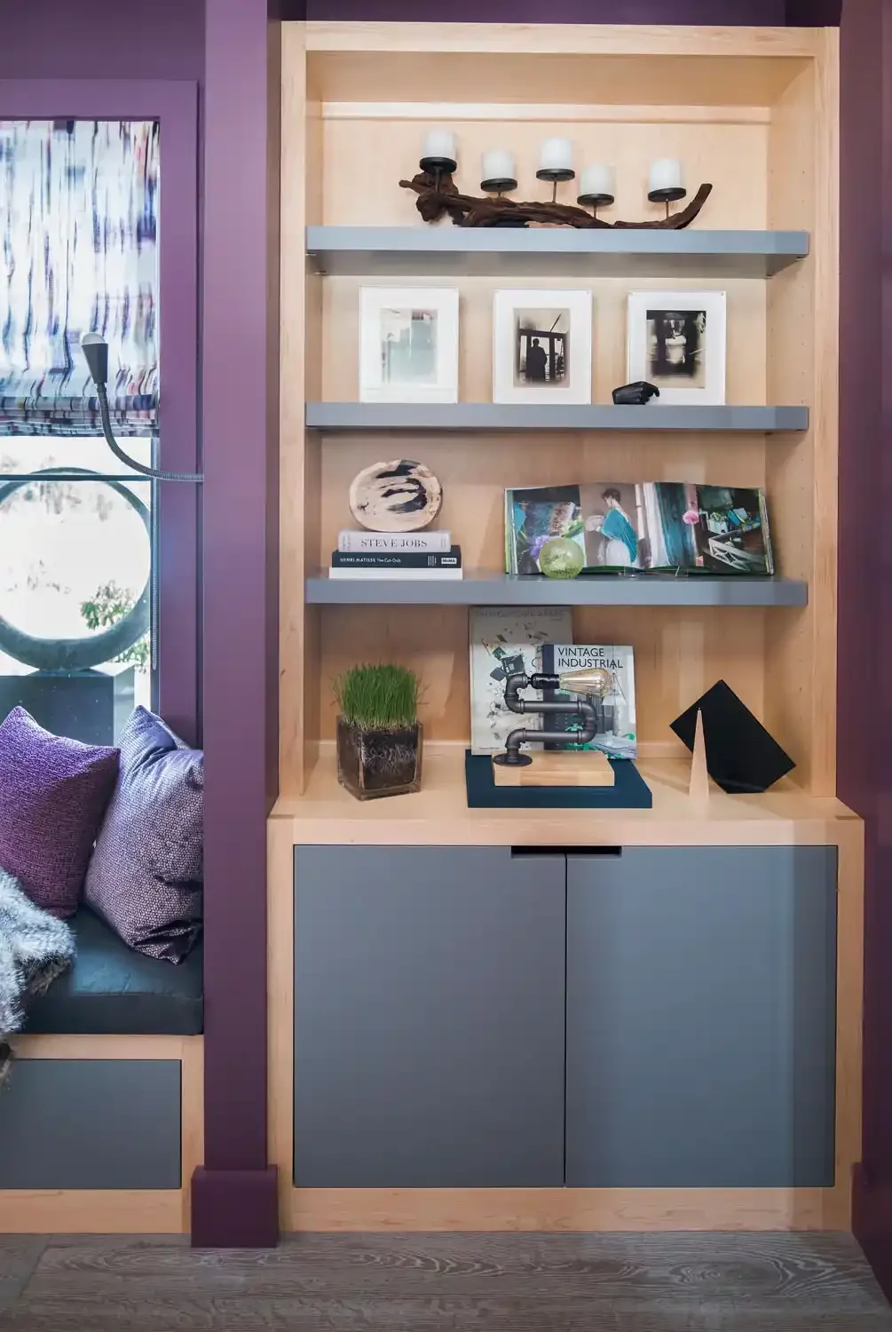 A built-in wooden shelf unit with three blue shelves holding decorative items, books, and framed photographs. The bottom area has a closed cabinet with blue doors, and a small indoor plant is placed on the left side of its surface. To the left, part 