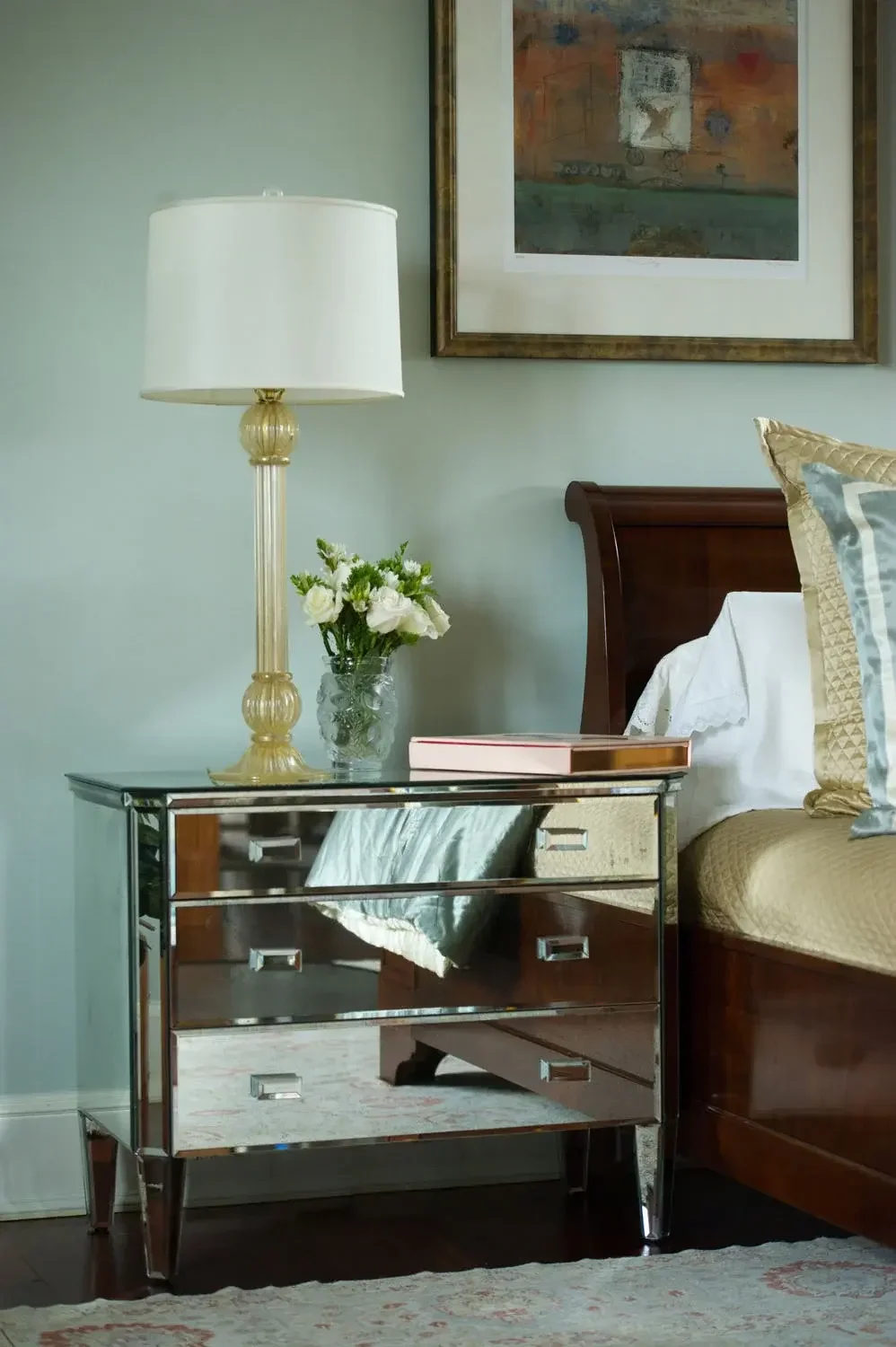 A bedroom nightstand with a mirrored finish, a table lamp with a white shade, a glass vase with white flowers, a hardcover book, and a decorative pillow. A framed abstract painting hangs on the wall above the bed with a wooden headboard, with part of