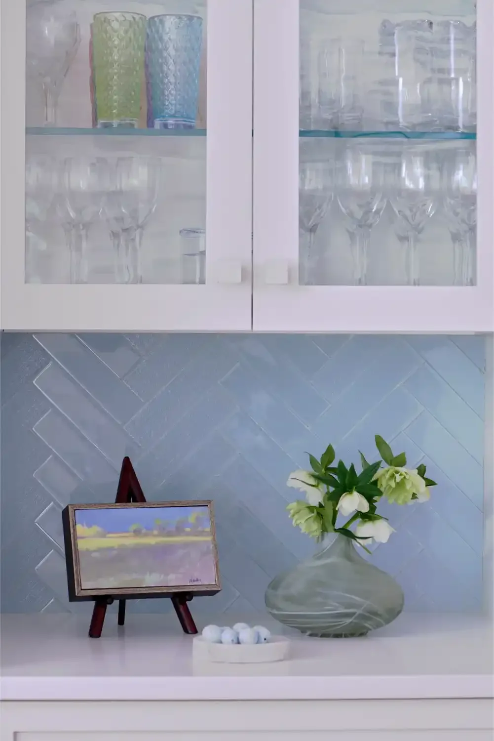 A white cabinet with glass doors containing colorful cups and clear glassware. Below, a white surface holds a small easel with a landscape painting, a large gray glass vase with white flowers, and a white dish with golf balls. The background features