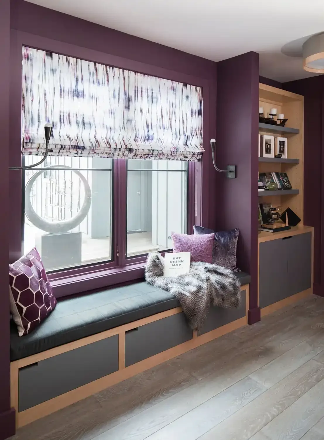 A window with a marble-like patterned Roman shade, a built-in window seat with cushions and pillows, a faux fur throw, and a small sign reading 'Eat Drink Nap' on the window seat. To the right is a built-in shelving unit with books, framed photos, an