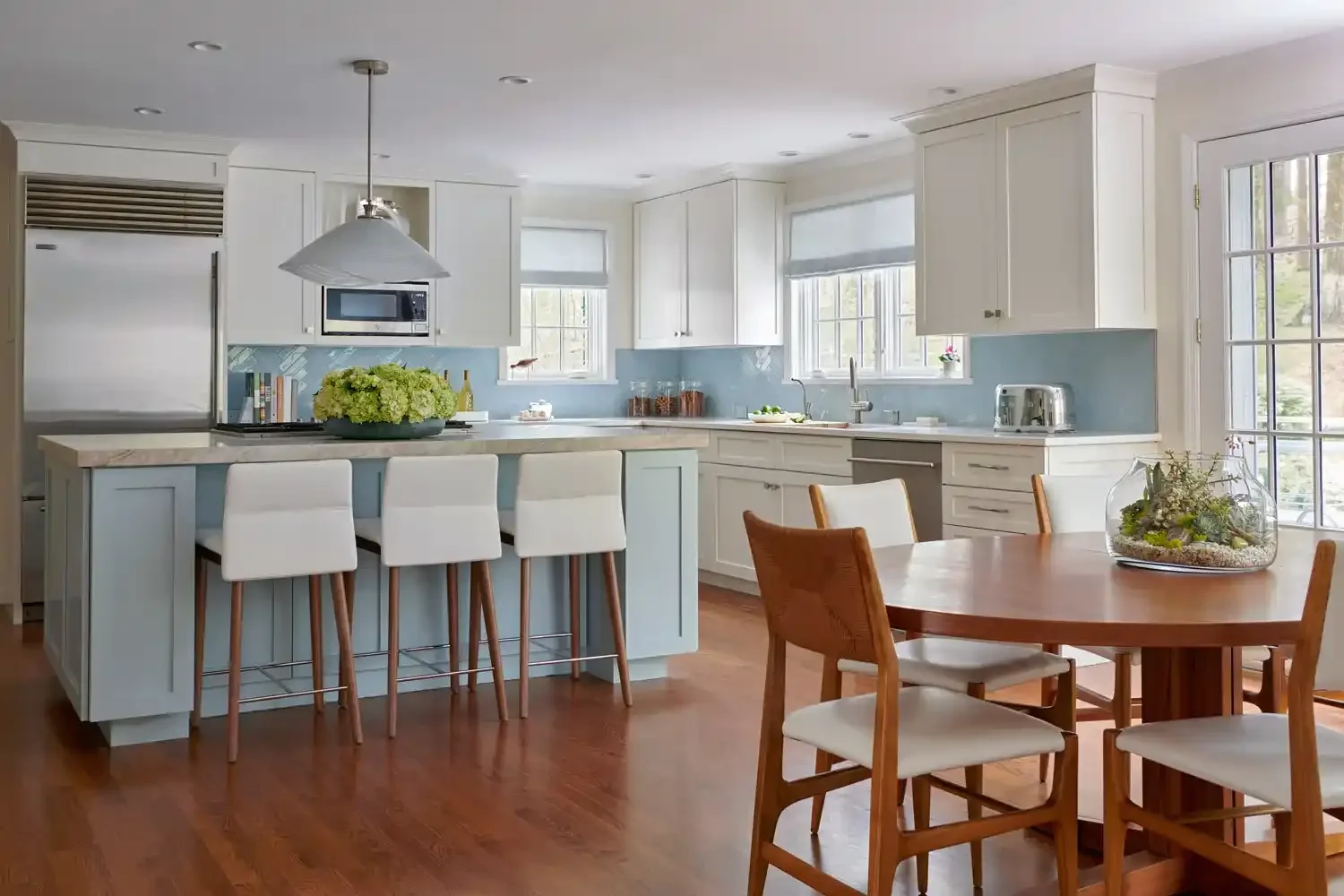 Bright kitchen with white cabinets, light blue backsplash, wooden dining table, and bar stools with white seats. Large windows let in natural light; decor includes a terrarium on the table and a floral centerpiece.