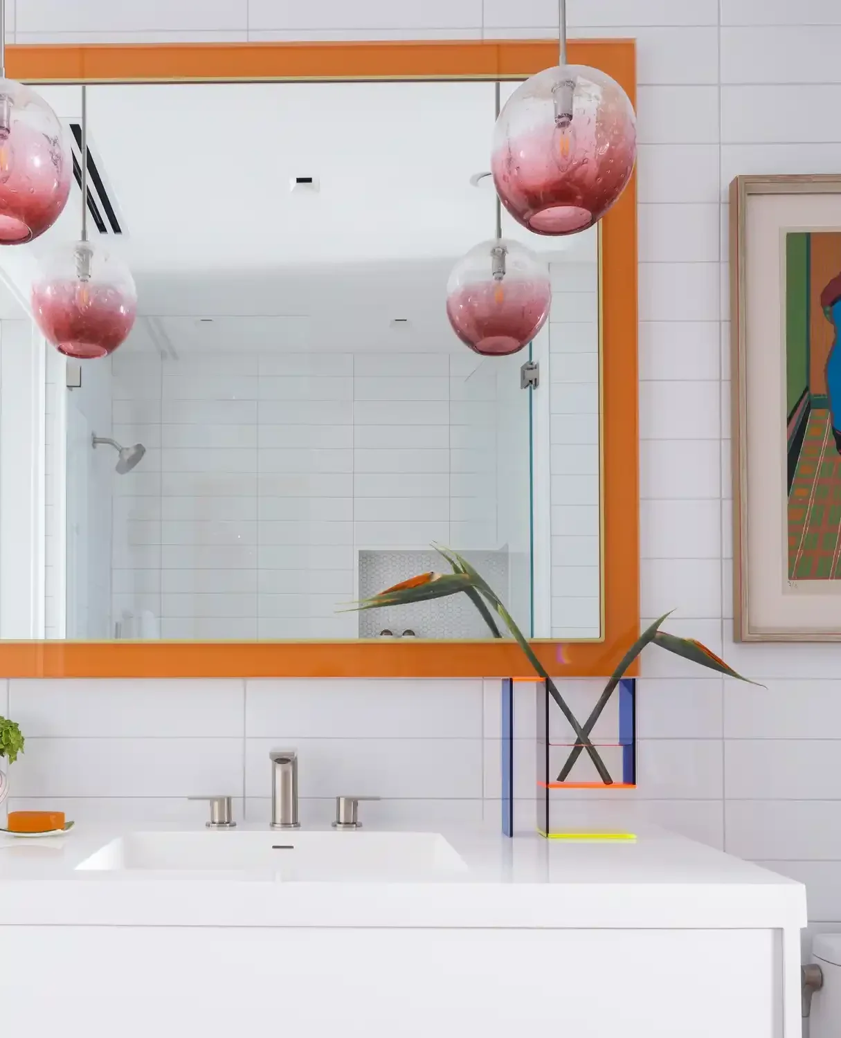 A bathroom mirror with an orange frame, three pink glass pendant lights hanging above, a white sink with a modern faucet, and a decorative plant with long leaves in a colorful geometric vase