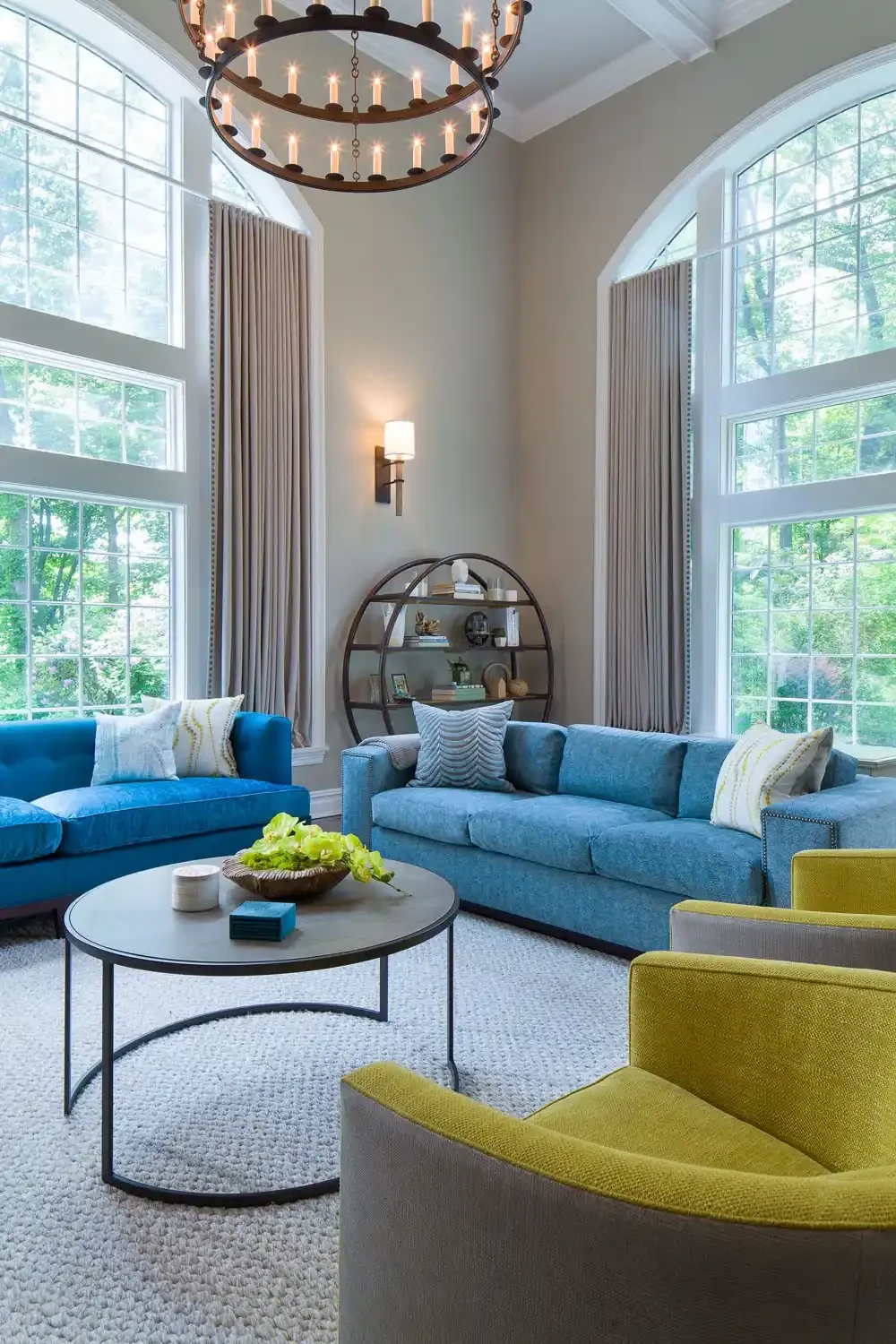 Living room with large arched windows, two blue sofas, a yellow armchair, a round coffee table with decorative items, a circular bookshelf, and a chandelier hanging from the ceiling.