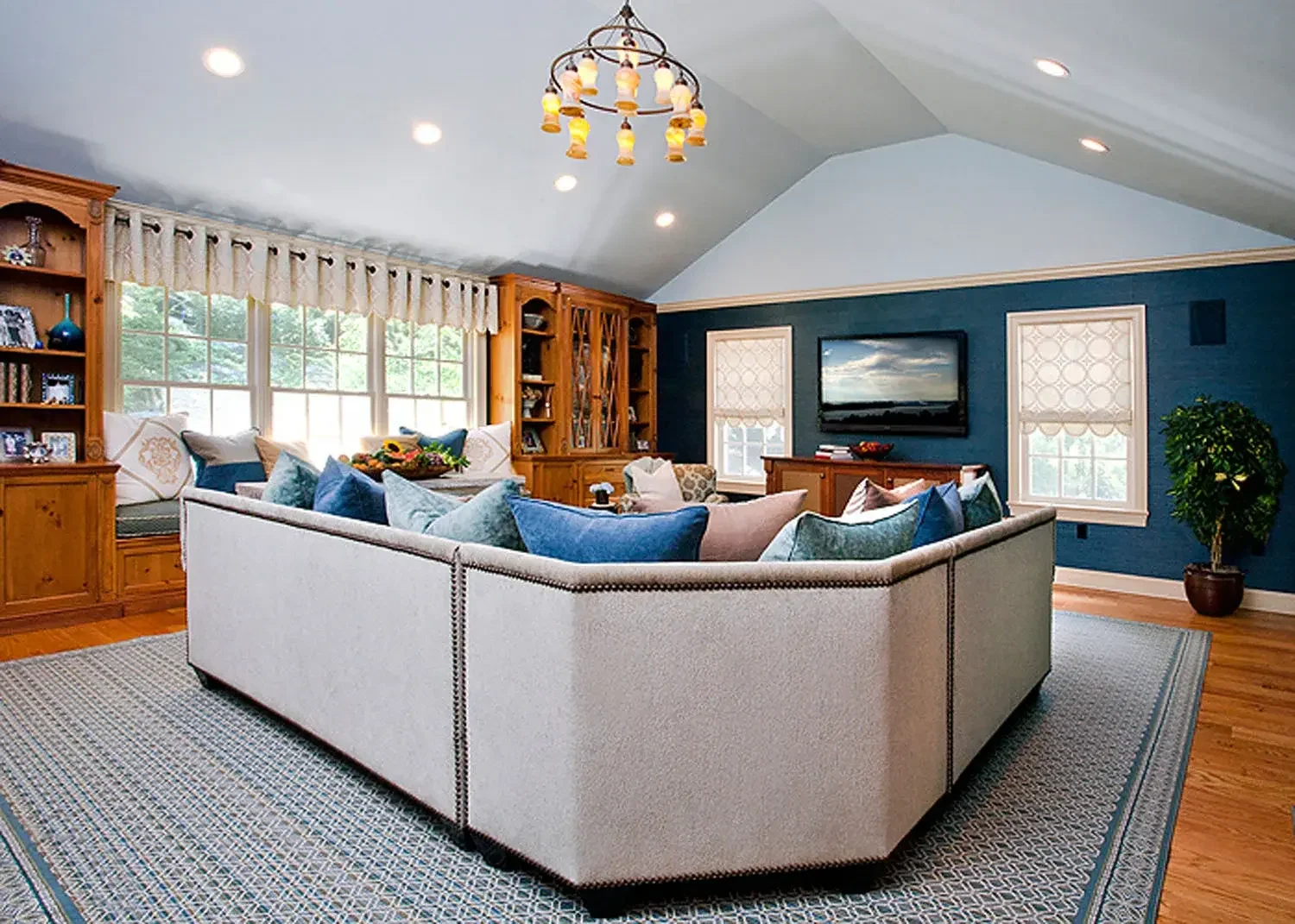 Bright living room with a large beige sectional sofa adorned with blue and gray pillows, wooden built-in shelves, a flat-screen TV on a blue accent wall, bay windows with valances, hardwood floors, a patterned area rug, and a hanging chandelier.