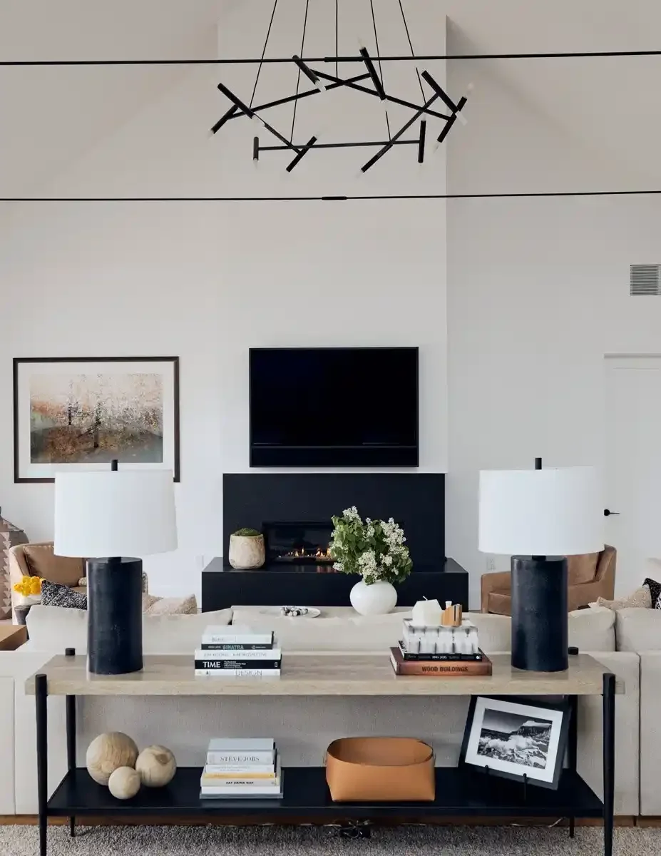Living room with white walls, a modern black chandelier, a flat-screen TV above a black fireplace, beige sofas, two white lamps on a wooden console table, decorative objects including a framed photo, books, a ceramic bowl, and a white vase with flowe