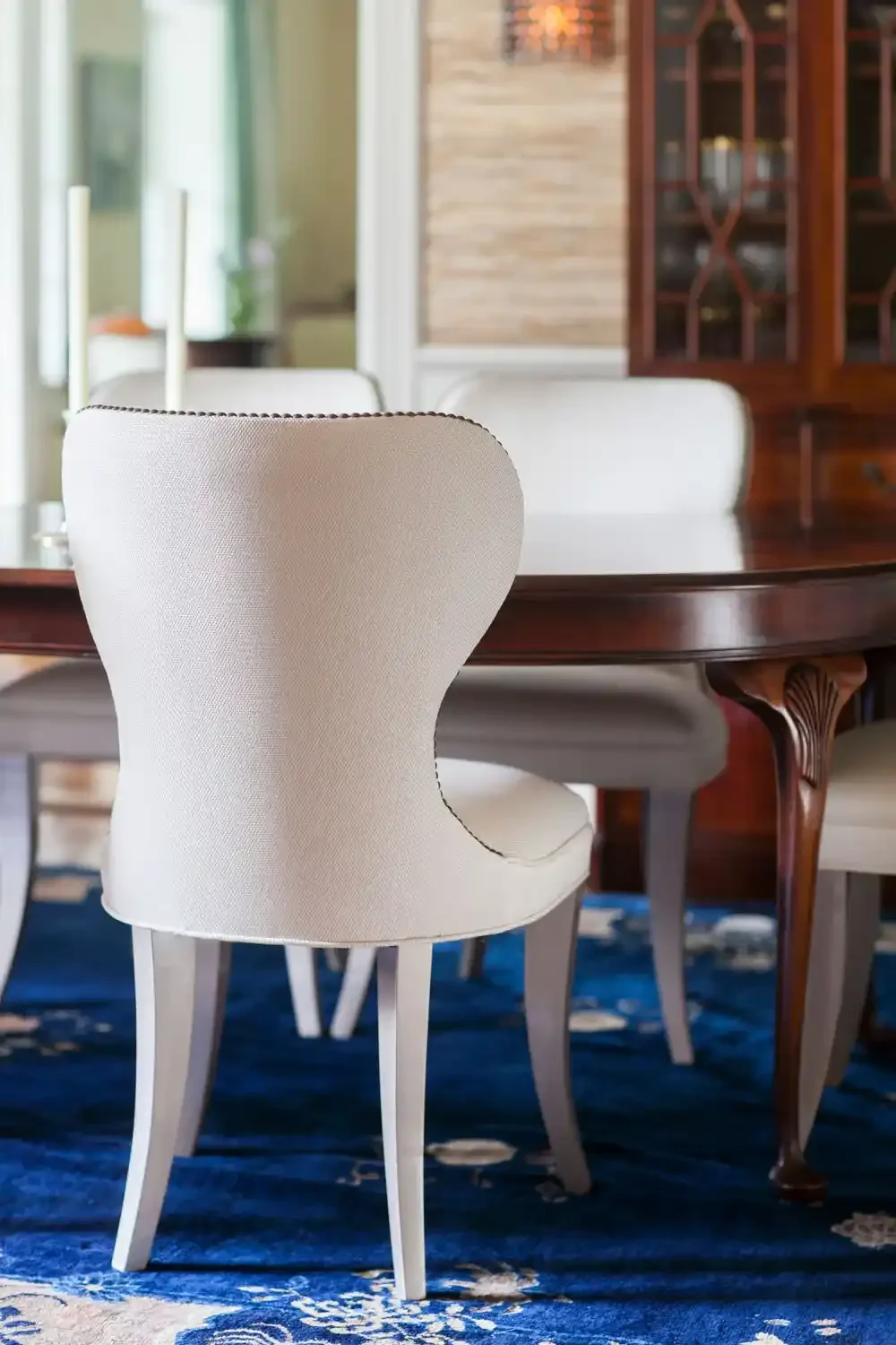 Close-up of a white upholstered dining chair with curved back, placed in front of a wooden dining table in a well-lit room.