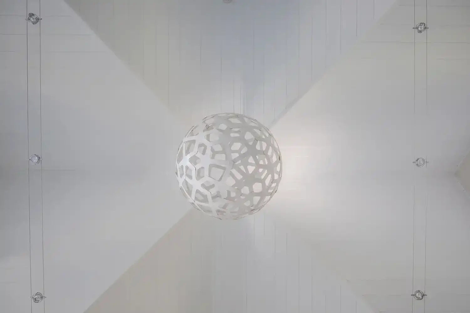 Modern white spherical pendant light with a geometric cutout pattern hanging from a white ceiling, illuminated and casting shadows.