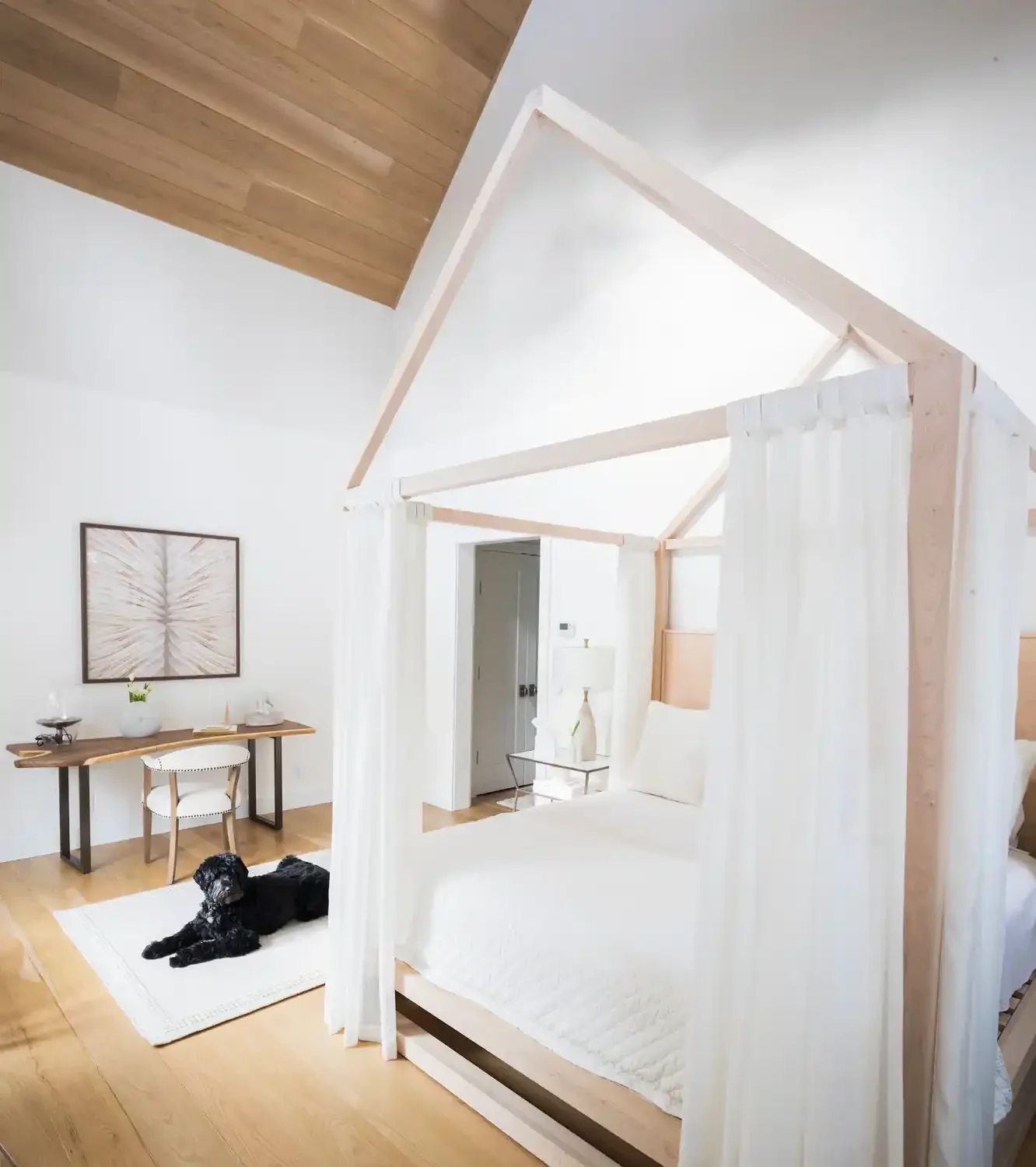 A bright, modern bedroom with a white canopy bed, white curtains, a wooden ceiling and hardwood floors. There is a black dog lying on a white rug, a small desk with a white chair, and a framed art piece on the wall.