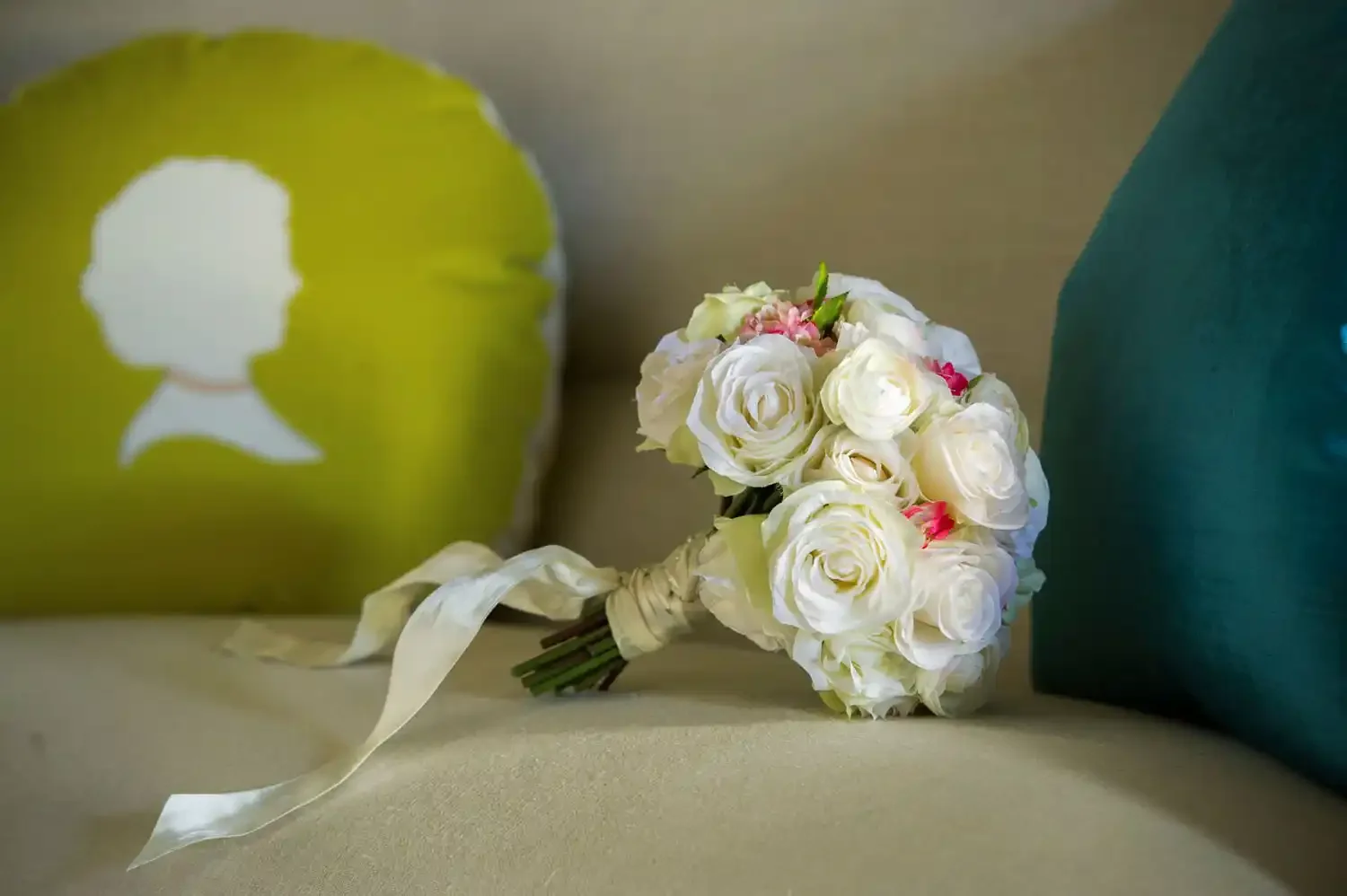 A bouquet of white roses with some pink accents, tied with a white ribbon, resting on a beige surface. Two balloons are visible in the background; one is green with a white silhouette of a woman's profile, and the other is not fully visible.