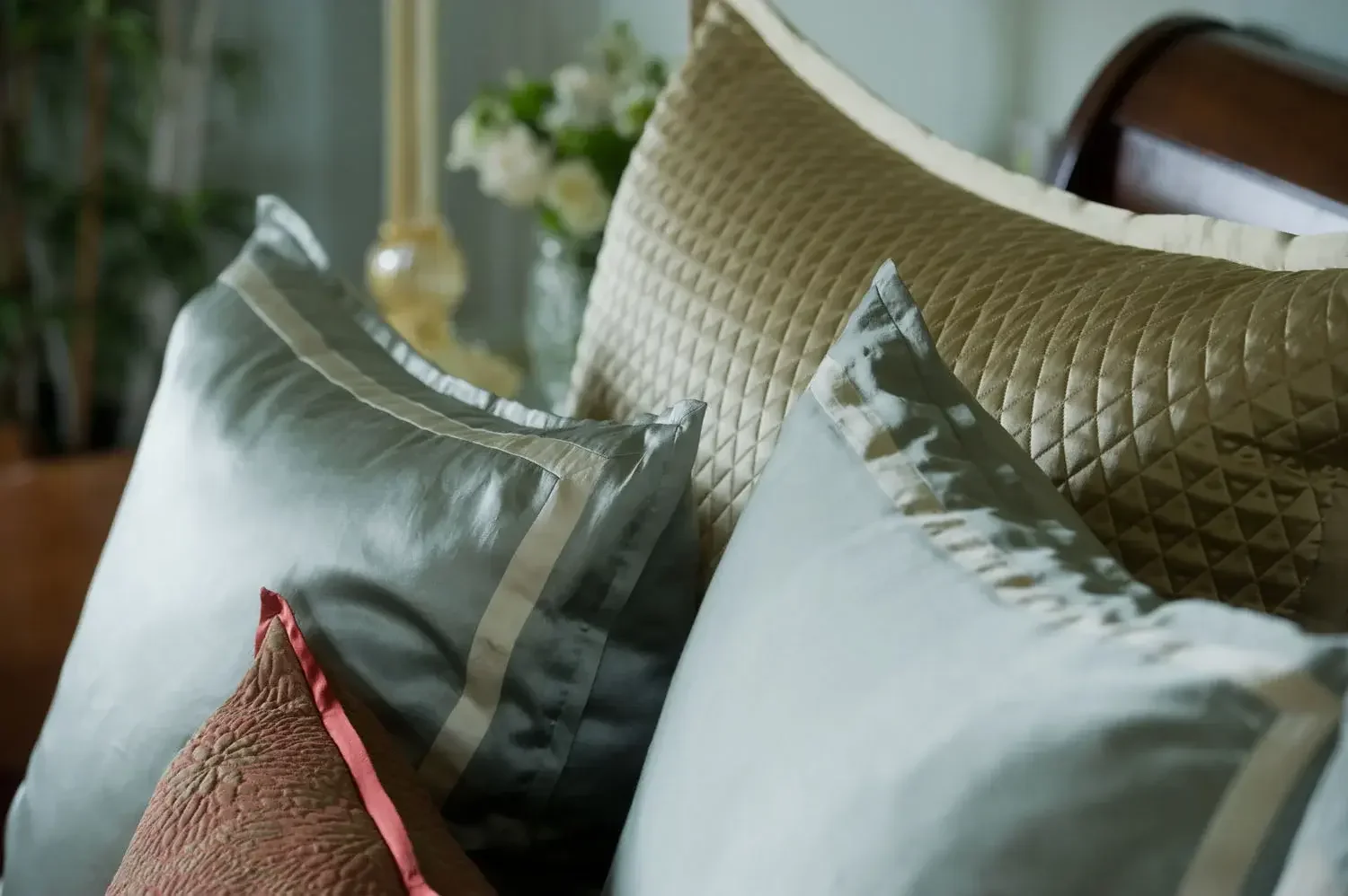 Close-up of decorative pillows on a bed, including one with a quilted beige pattern, one with a metallic sheen, and another with a rust color and pink piping.