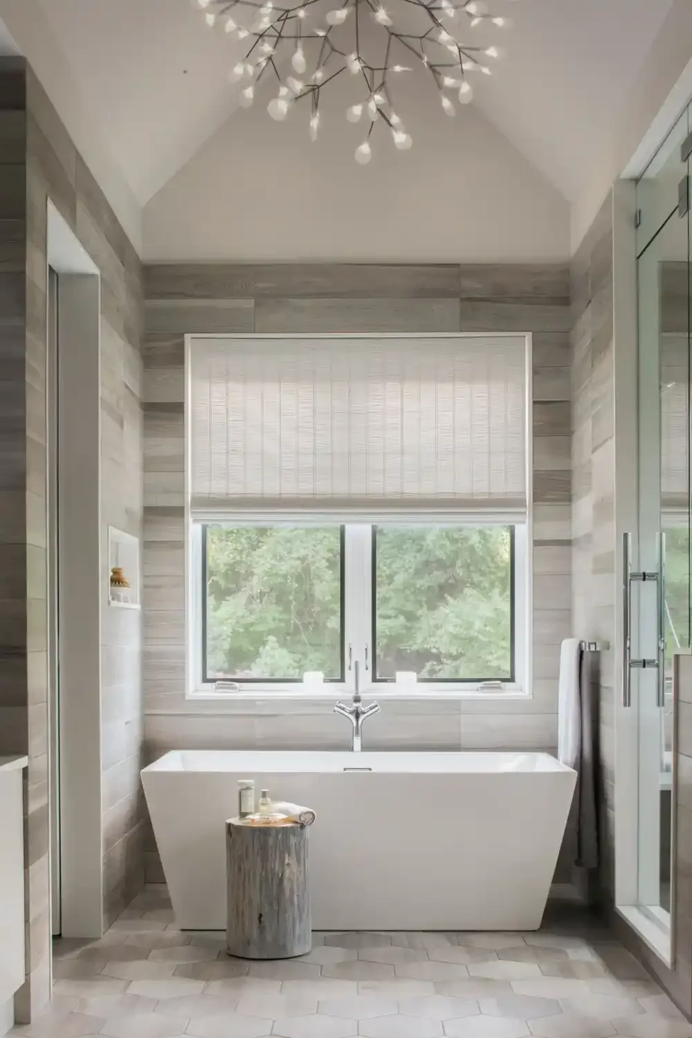 Modern bathroom featuring a white bathtub beneath a window with a view of green trees, a chandelier on the ceiling, and light wood wall panels.