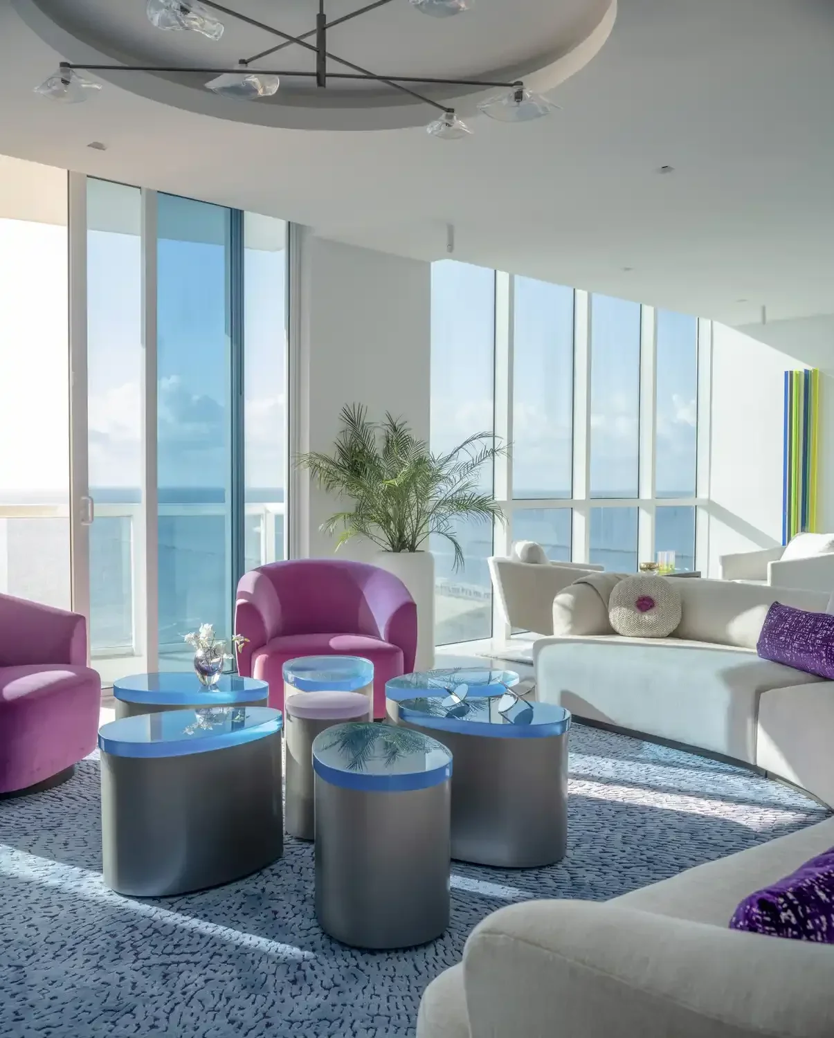 Modern living room with white and purple furniture, glass tables, large windows showing a view of the sky and ocean, and a potted plant.