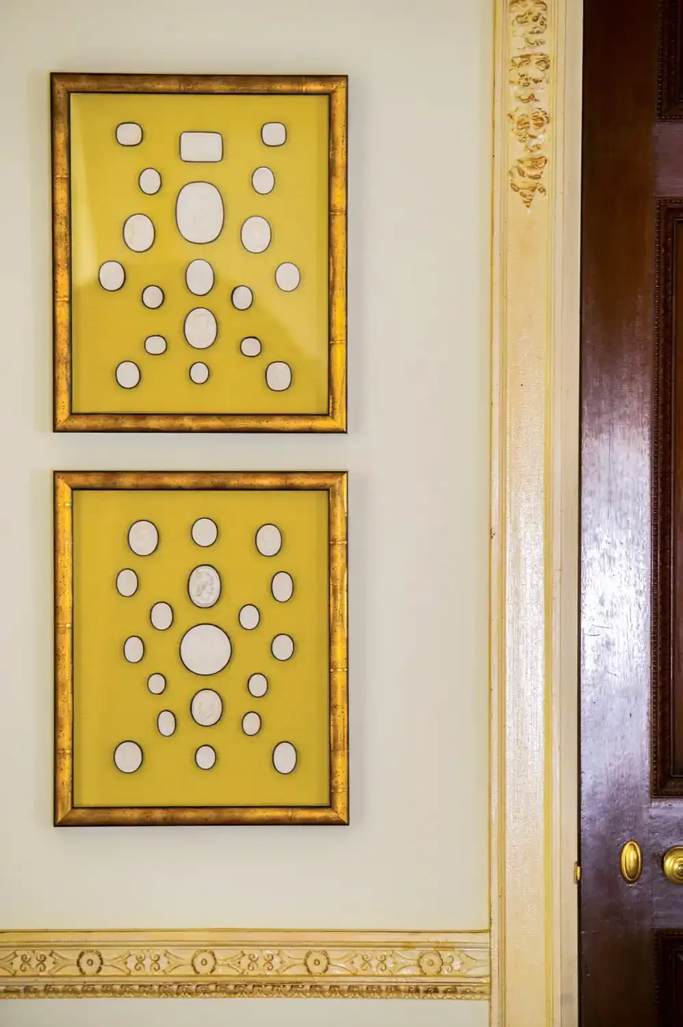 Two framed abstract art pieces with yellow backgrounds and white oval and circle shapes, hung vertically on a light-colored wall with ornate gold trim and a decorative border.