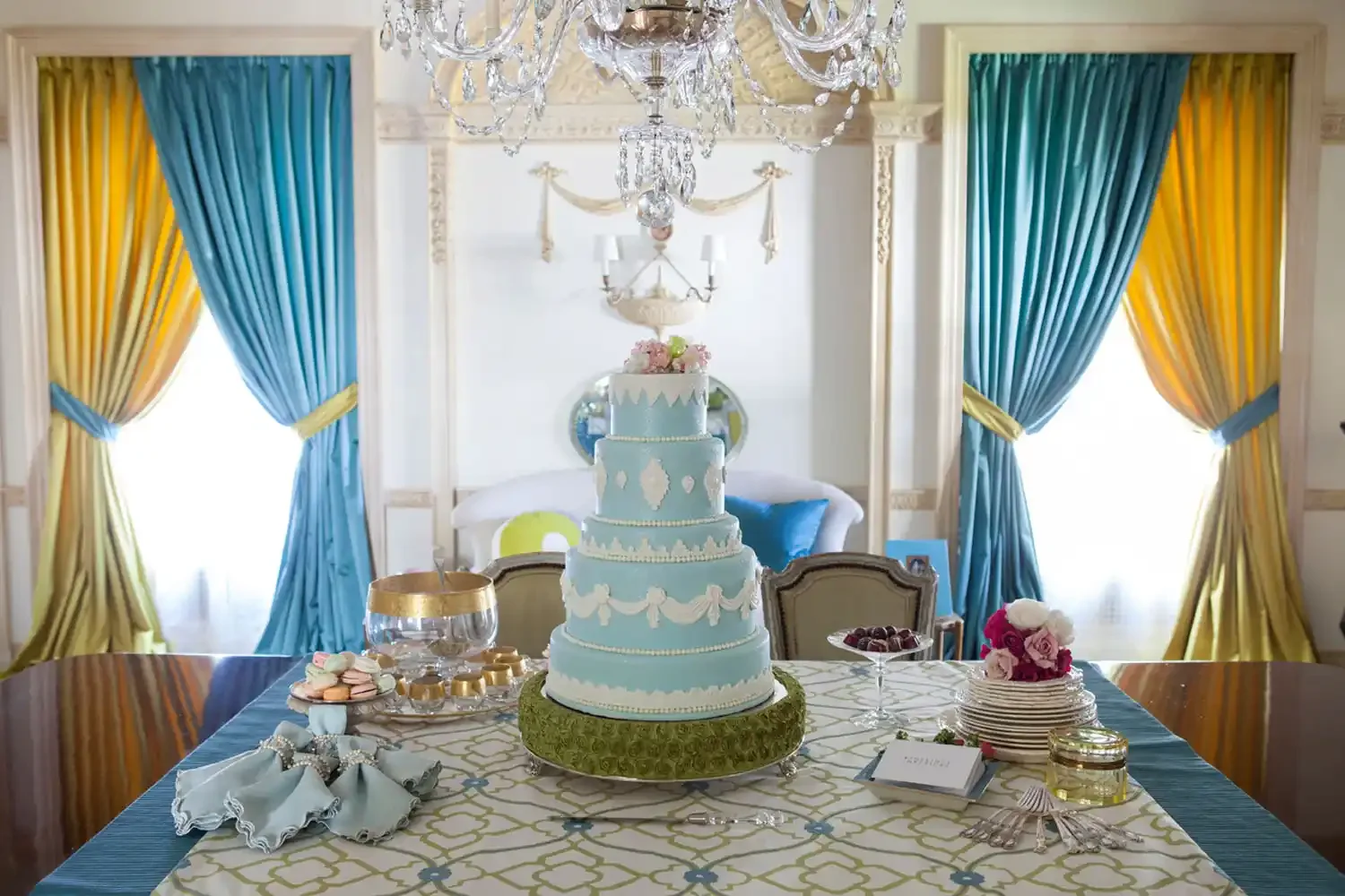 Elegant dining room decorated for a celebration with a multi-tiered blue and white cake on the table, surrounded by sweets, flowers, and festive decorations. The room features yellow and teal curtains, a crystal chandelier, and classic furniture.