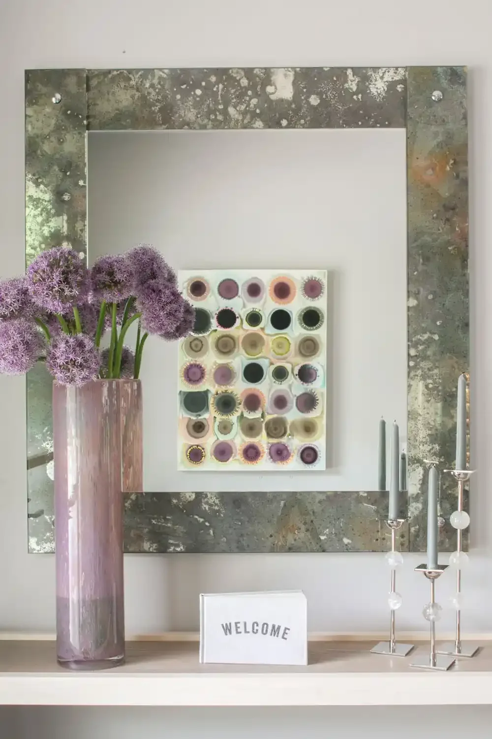 Decorative shelf with a tall pink vase holding purple allium flowers, a white sign with 'WELCOME' in black letters, three modern candlestick holders with white candles, and a colorful wall art piece with circular patterns, all set against a mirror wi