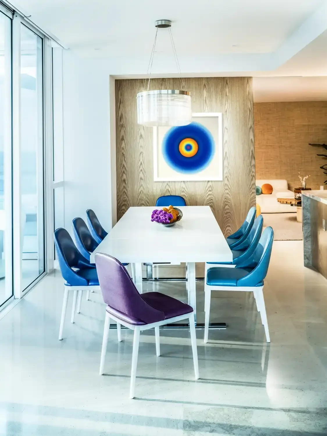 Dining room with white table, eight chairs with blue and purple upholstery, and abstract circular wall art.