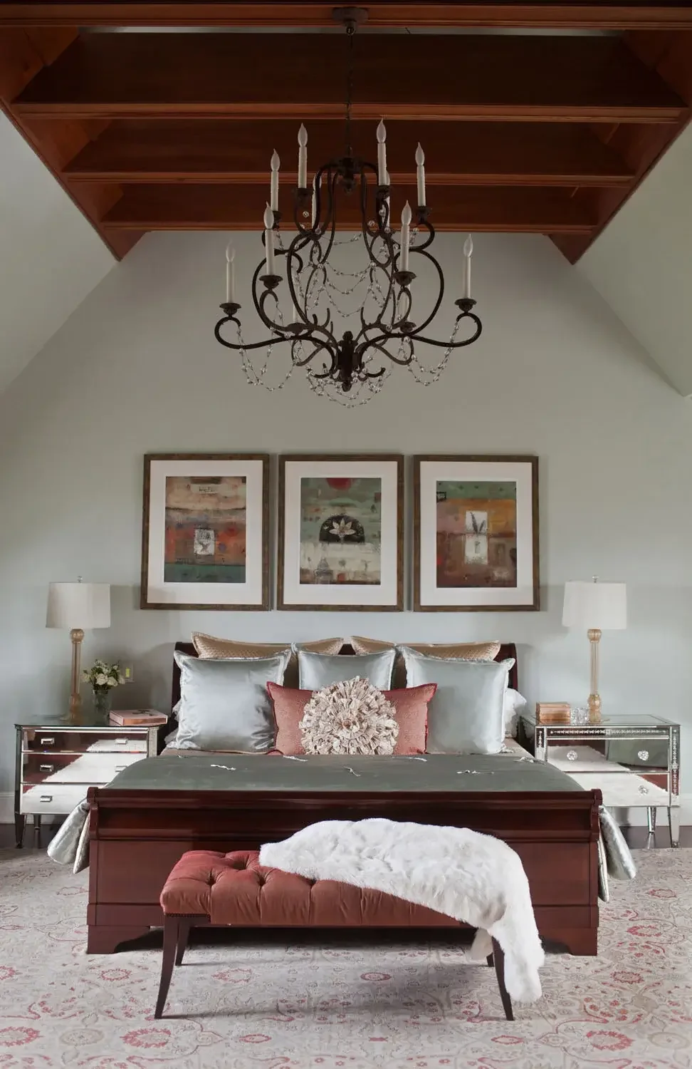 Elegant bedroom with a wooden bed, decorative pillows, mirrors, and framed artwork. A luxurious chandelier hangs from a high ceiling, and a tufted bench with a white throw covers the foot of the bed. Lamps and flowers are on mirrored nightstands.