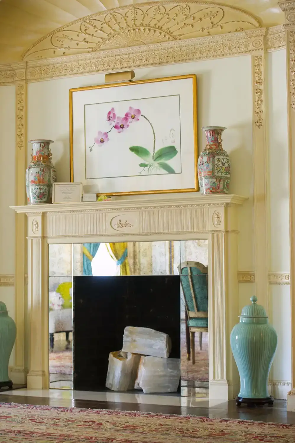 Elegant living room with ornate white fireplace mantle, pink orchid artwork above, two large colorful vases on the mantle, a framed mirror in the fireplace opening reflecting a room with chairs and curtains, and a large ginger jar on the right side.