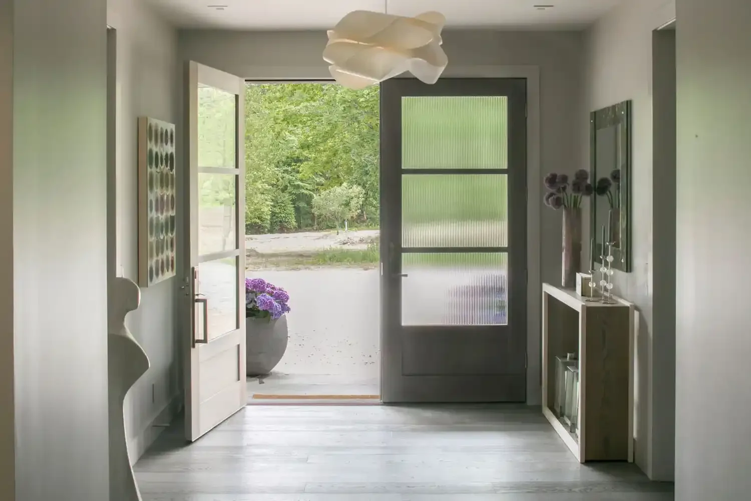 View of a modern entryway with an open door leading outside to a greenery landscape, including potted purple flowers, artwork, and decorative items inside.