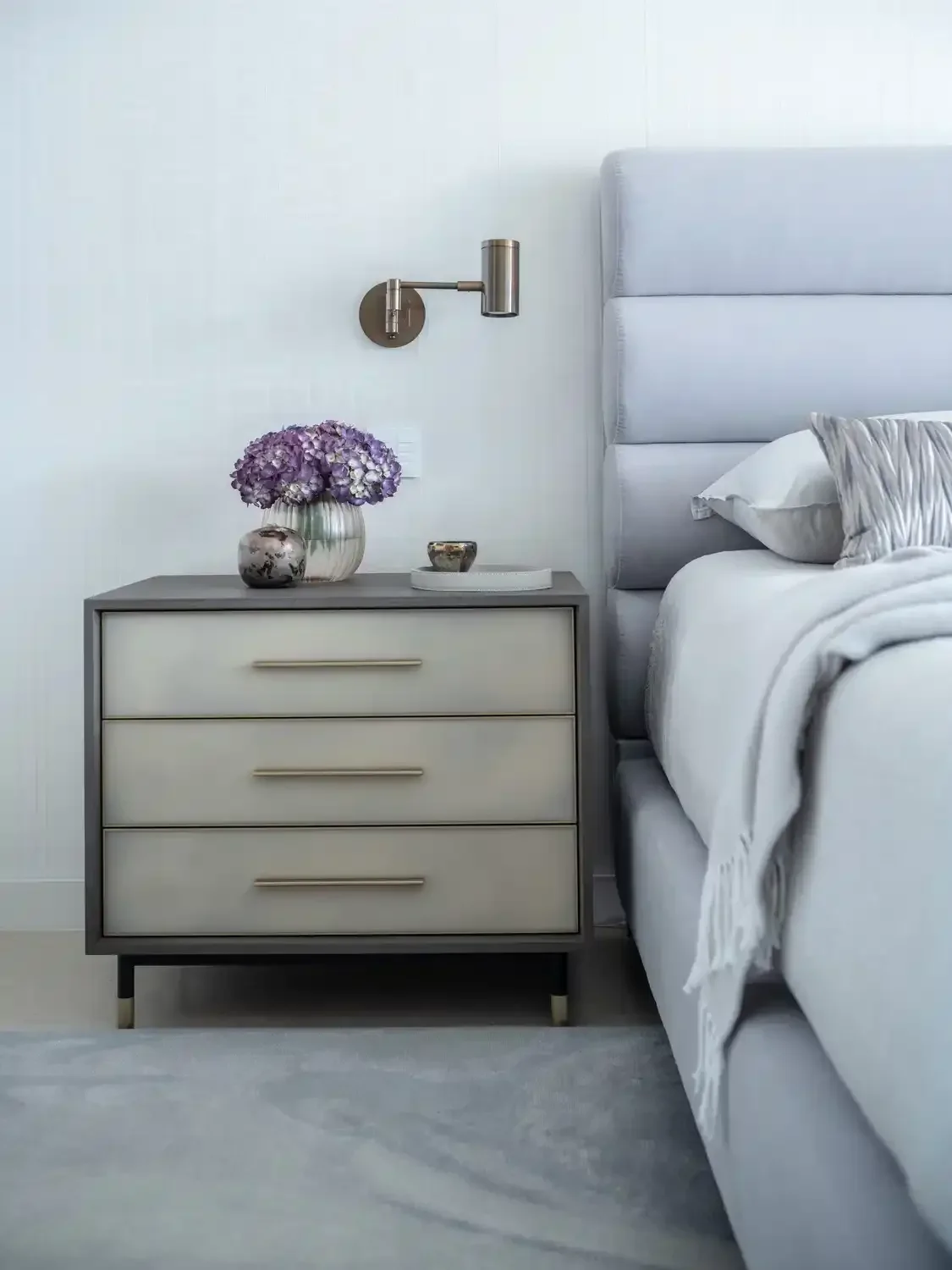 A nightstand next to a bed with a grey upholstered headboard. The nightstand has three drawers with horizontal handles. On top of the nightstand are two vases with purple flowers and decorative items. A wall-mounted reading light is above the nightst
