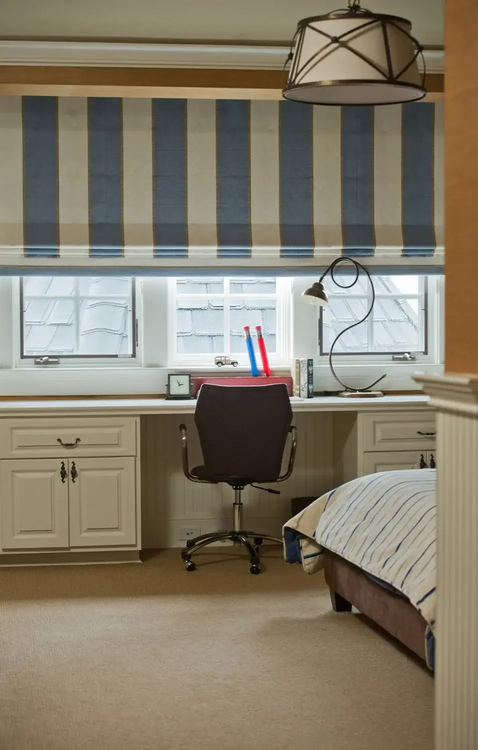 A bedroom workspace with a desk by two windows, a black office chair, a small desk clock, a desk lamp, two large colored pencils, books, and a striped bed partially visible on the right, with a beige carpeted floor.