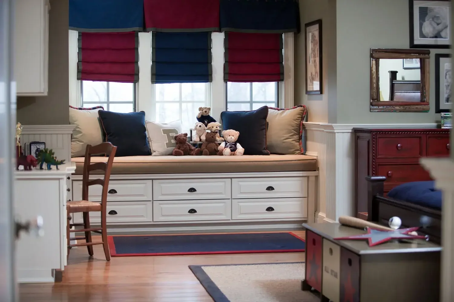 A cozy window seat with stuffed teddy bears and large cushions, surrounded by framed artwork and a mirror in a well-lit room with hardwood floors and a multicolored area rug.