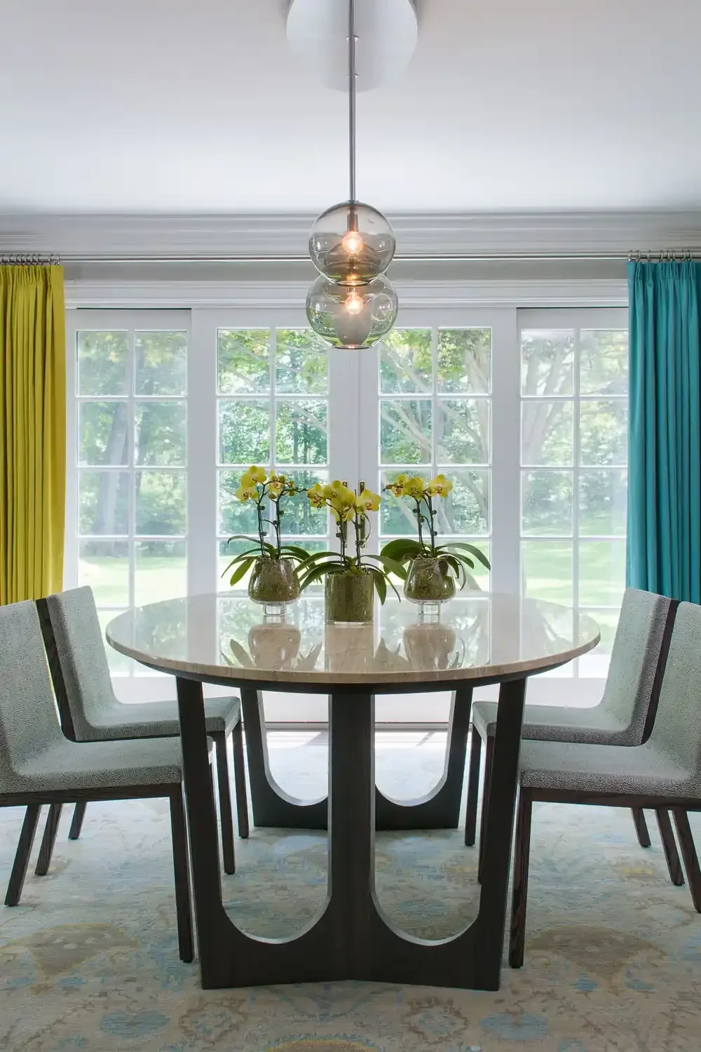 A round dining table with four gray upholstered chairs around it, three potted yellow orchids in the center, large windows with green trees outside, and colorful yellow and blue curtains, with a modern light fixture hanging above.