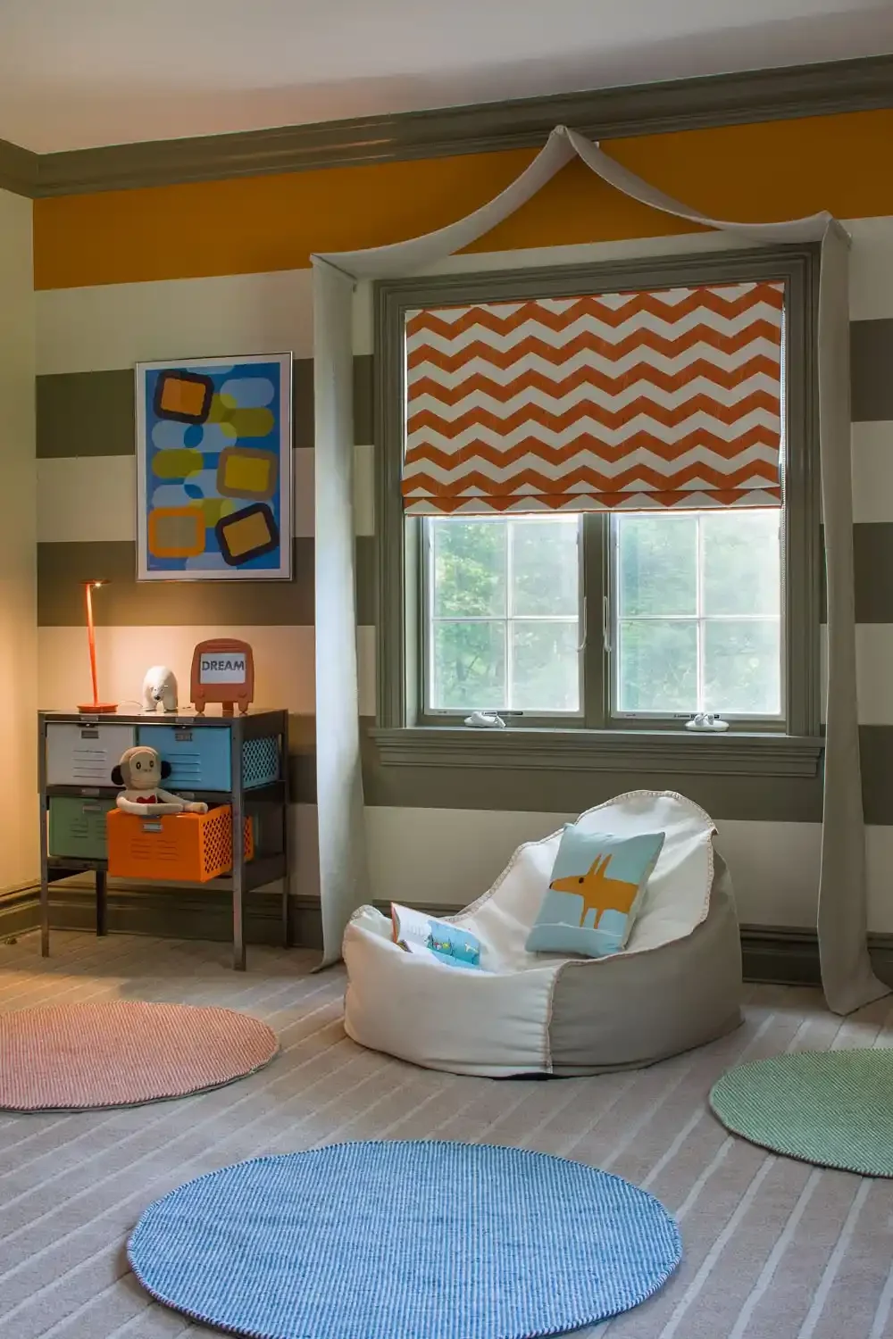 A cozy children's room with a window covered by an orange and white chevron roller blind, featuring colorful oval wall art, a white bean bag chair with a yellow fox pillow, a small shelving unit with toys and a lamp, and three circular rugs in pink, 