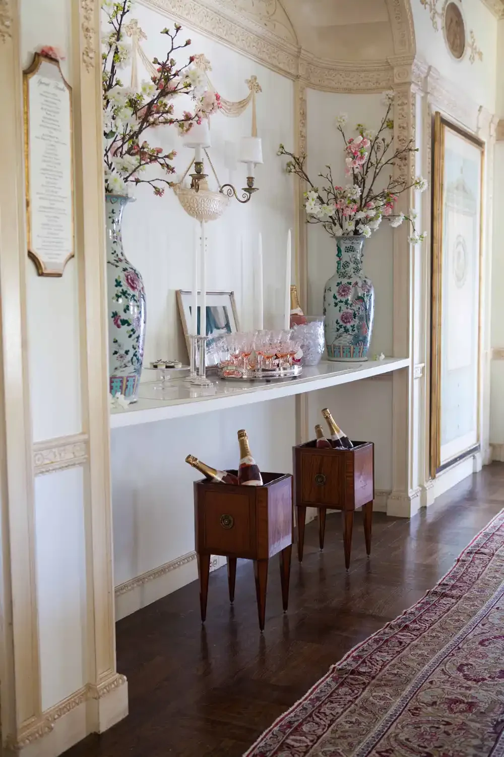 Elegant sideboard with large porcelain vases filled with cherry blossom branches, framed pictures, glasses of pink champagne, and small champagne bottles on wooden stands, in a richly decorated room with ornate moldings and a Persian rug.