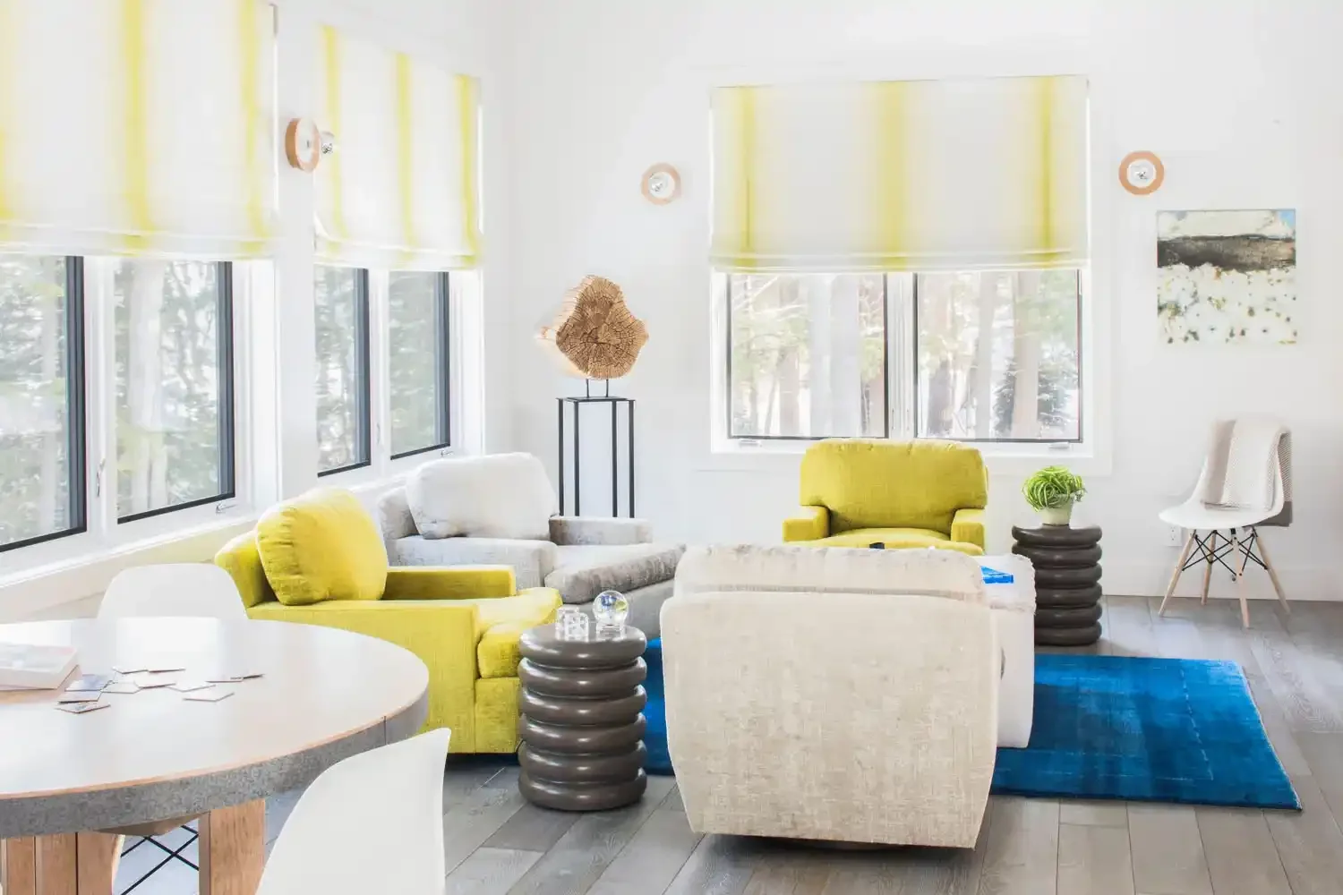 Bright living room with yellow and white seating, large windows with yellow blinds, blue rug, and modern decor.
