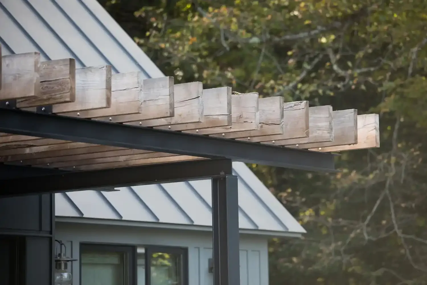 Close-up view of the edge of a wooden deck or platform under construction, with visible support beams and metal framing, adjacent to a modern house with gray siding and a window, set against a background of trees.