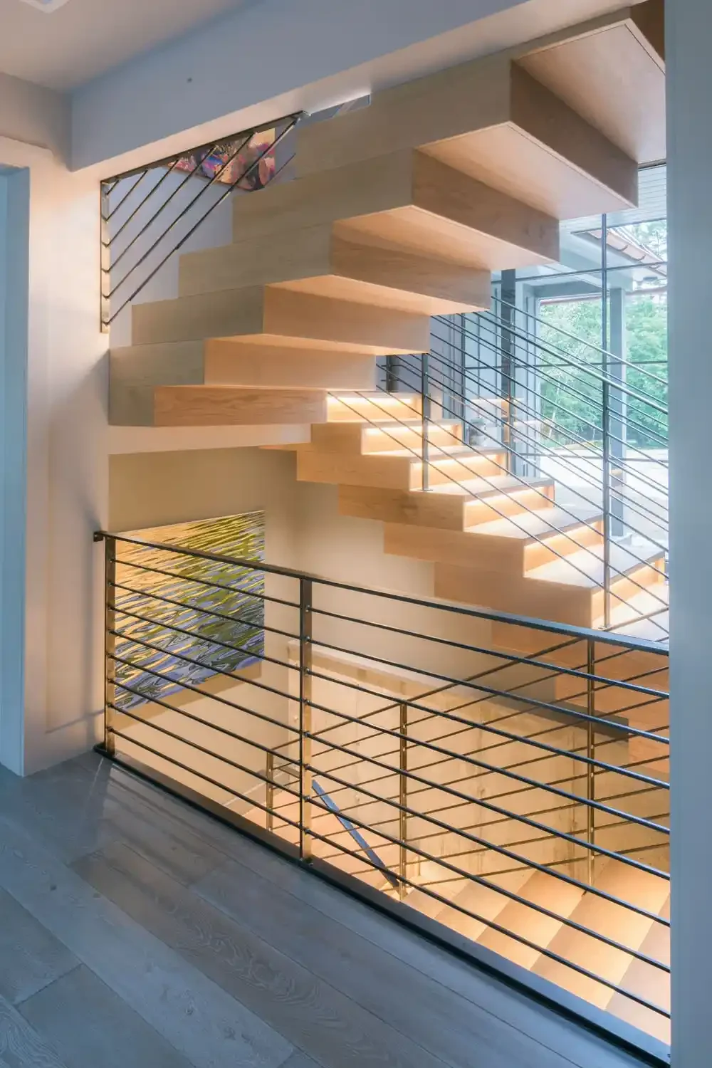 Modern wooden staircase with metal railings in a well-lit room with large windows and a colorful abstract painting on the wall.