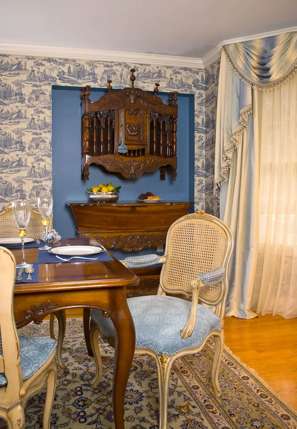 A dining room with a wooden table, ornate chairs, blue and cream curtains, vintage wallpaper, and a carved wooden hutch with decorative items on top.