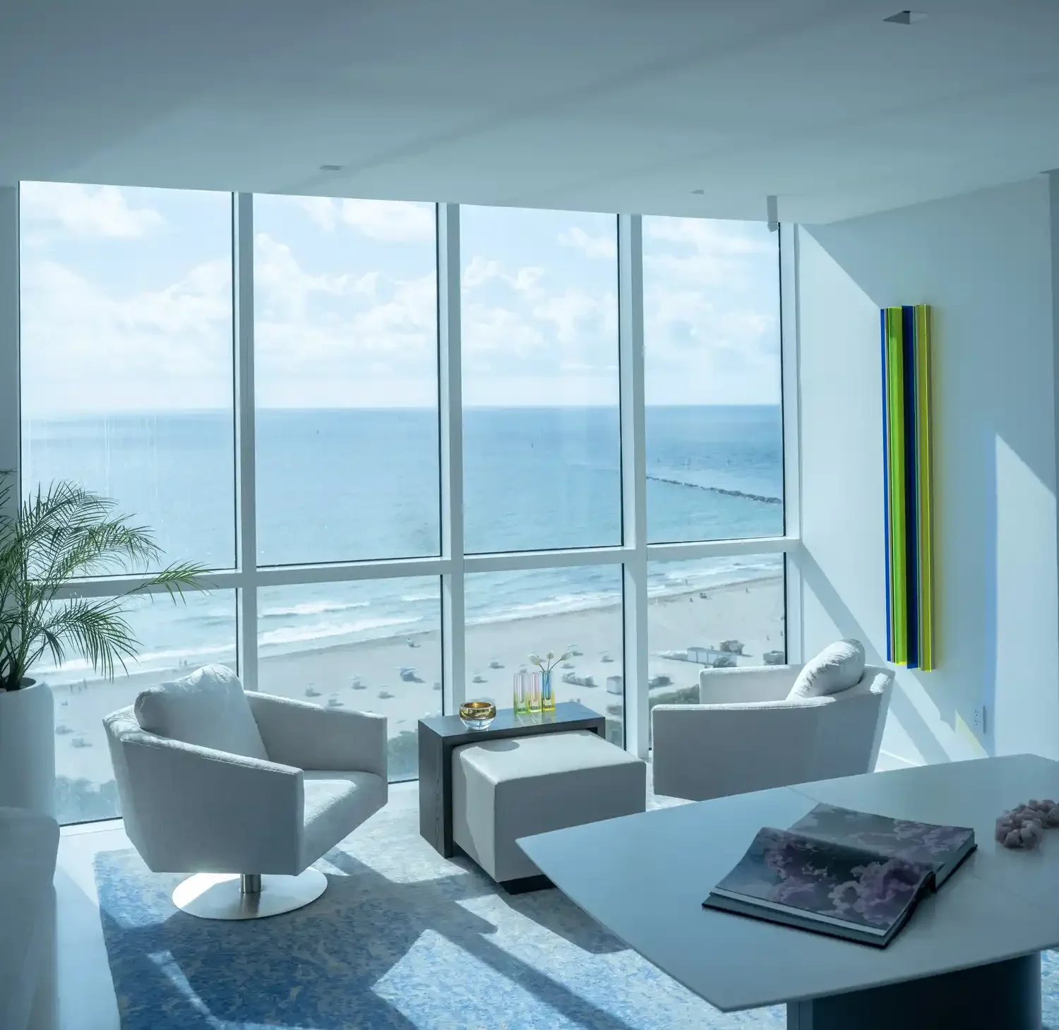 Modern living room with large floor-to-ceiling windows overlooking the beach and ocean, white chairs, a small table with magazines, and colorful abstract wall art.