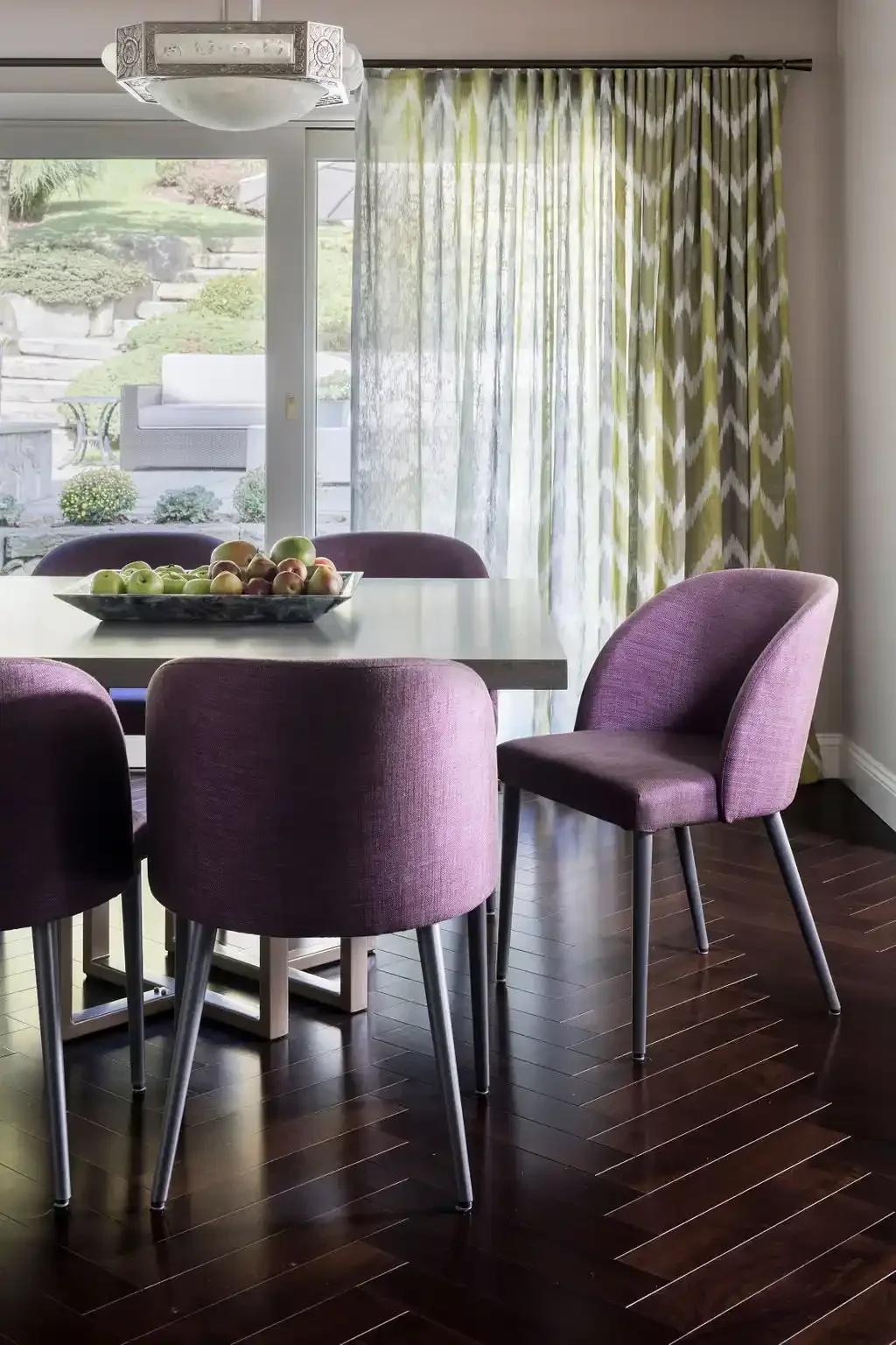 A dining room with a rectangular white table, purple upholstered chairs, a bowl of apples, and large windows with patterned curtains overlooking an outdoor patio and garden.