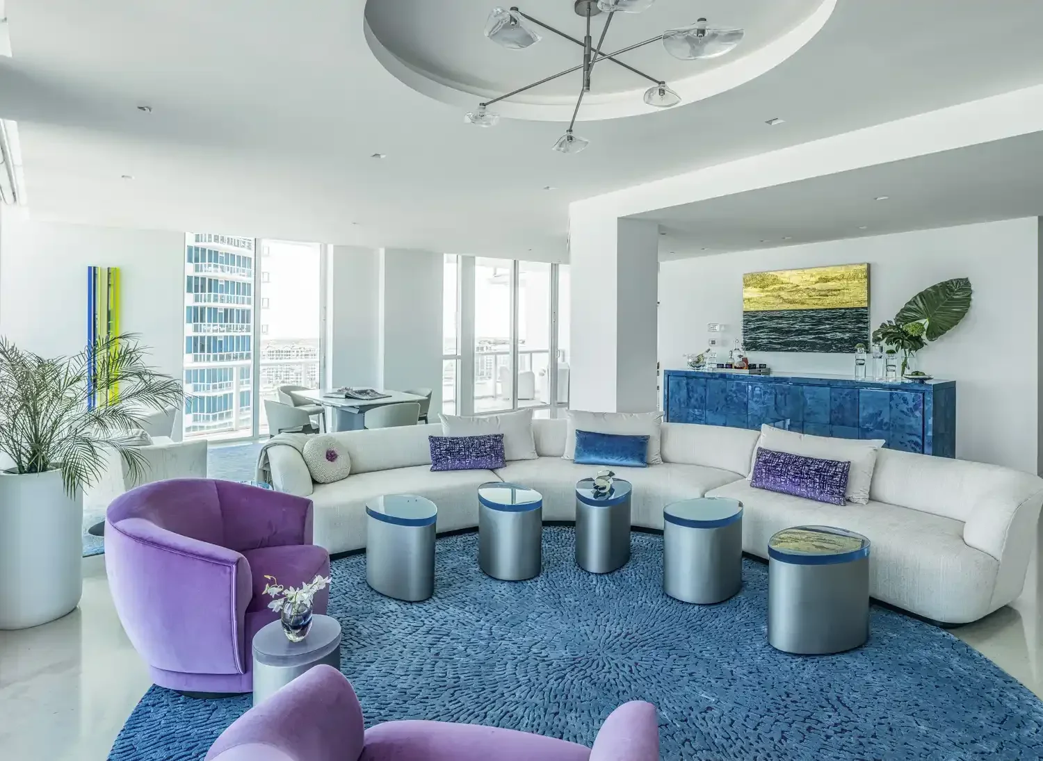 Modern living room with a white L-shaped sofa, purple velvet armchairs, and a blue area rug. Large windows with city views, a wall-mounted TV above a blue console, and a dining area in the background.