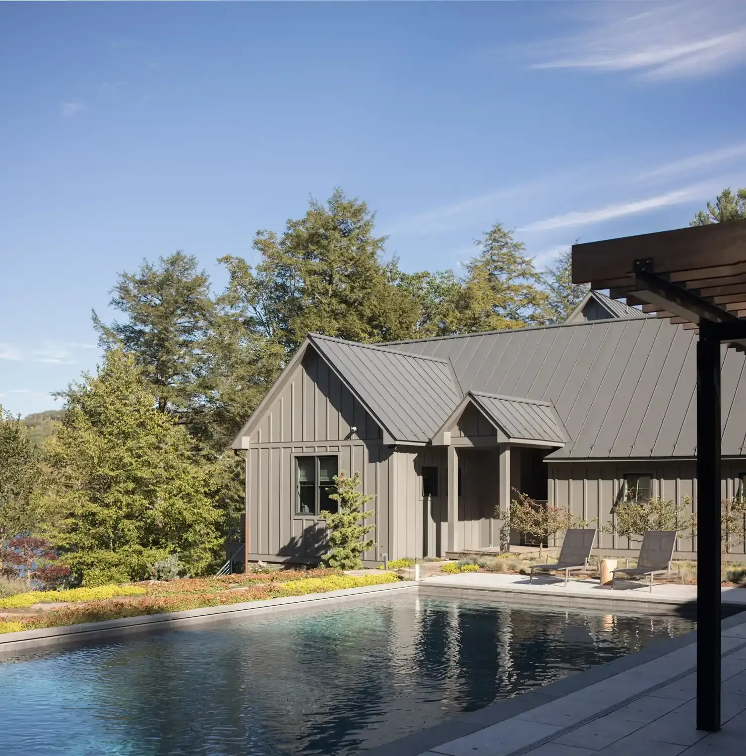 Modern house with gray exterior and metal roof next to a swimming pool, surrounded by trees and outdoor seating under a clear blue sky.