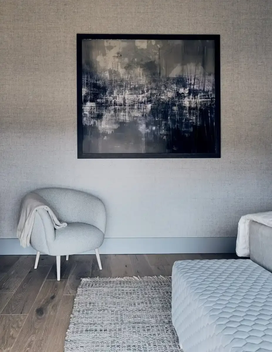 Modern living room with a light gray armchair, a black-framed abstract painting on the wall, a gray textured area rug, and part of a bed with a quilted cover.