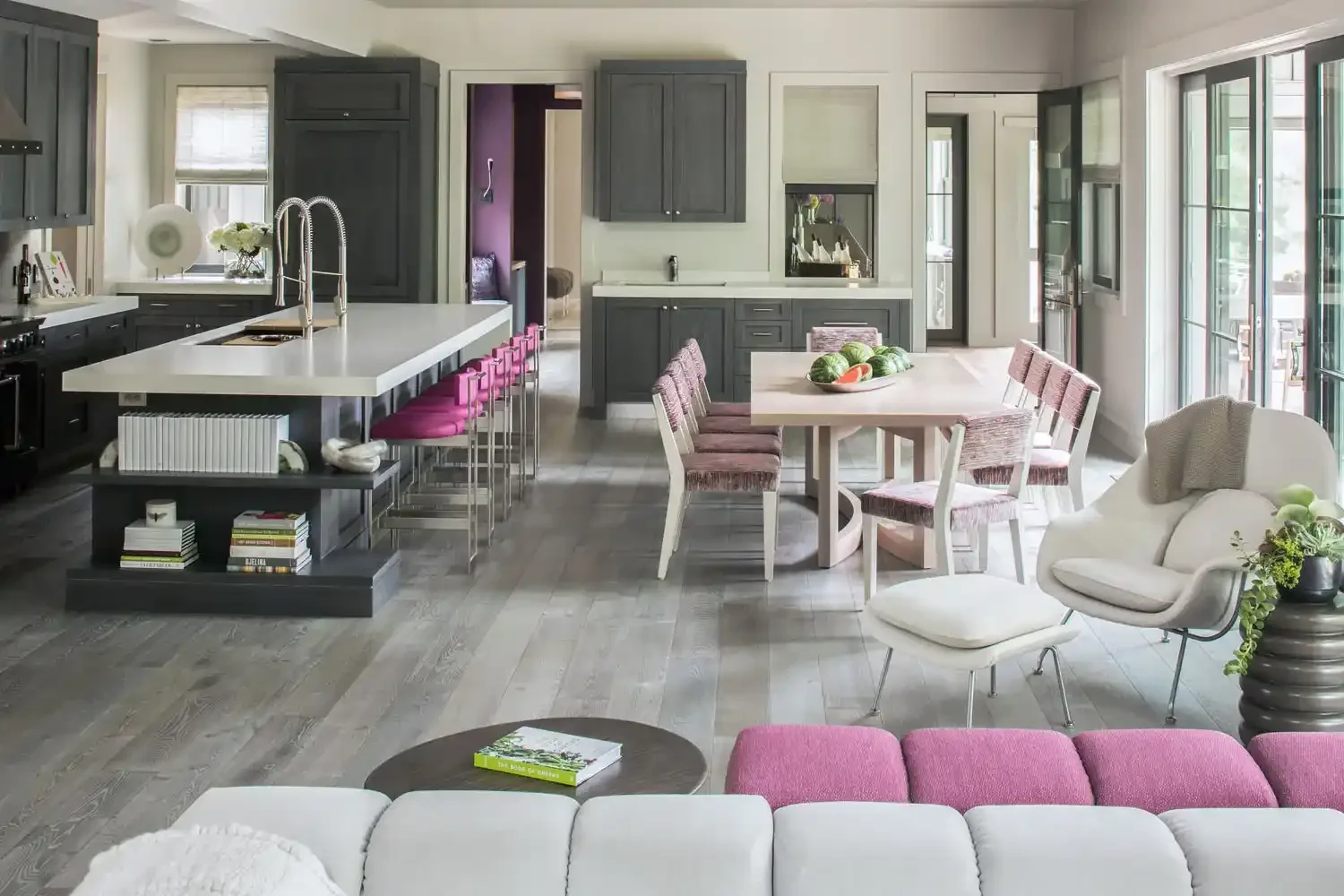 Open-concept kitchen and dining area with gray cabinets, white countertops, pink upholstered bar stools, and a light wood dining table with pink chairs, adjacent to a living space with white and pink furniture, large windows, and hardwood floors.