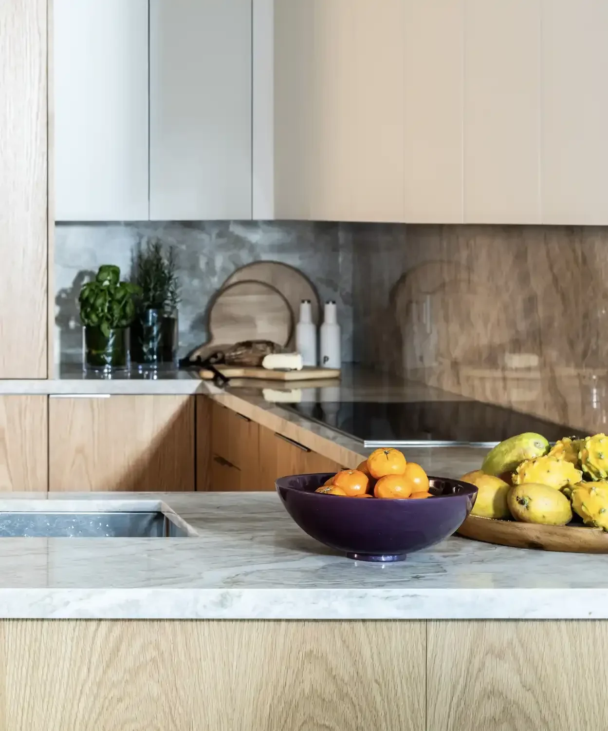 Modern kitchen with beige cabinetry, a marble counter, bowls of persimmons and green papayas, and kitchen utensils and potted herbs on the countertop.