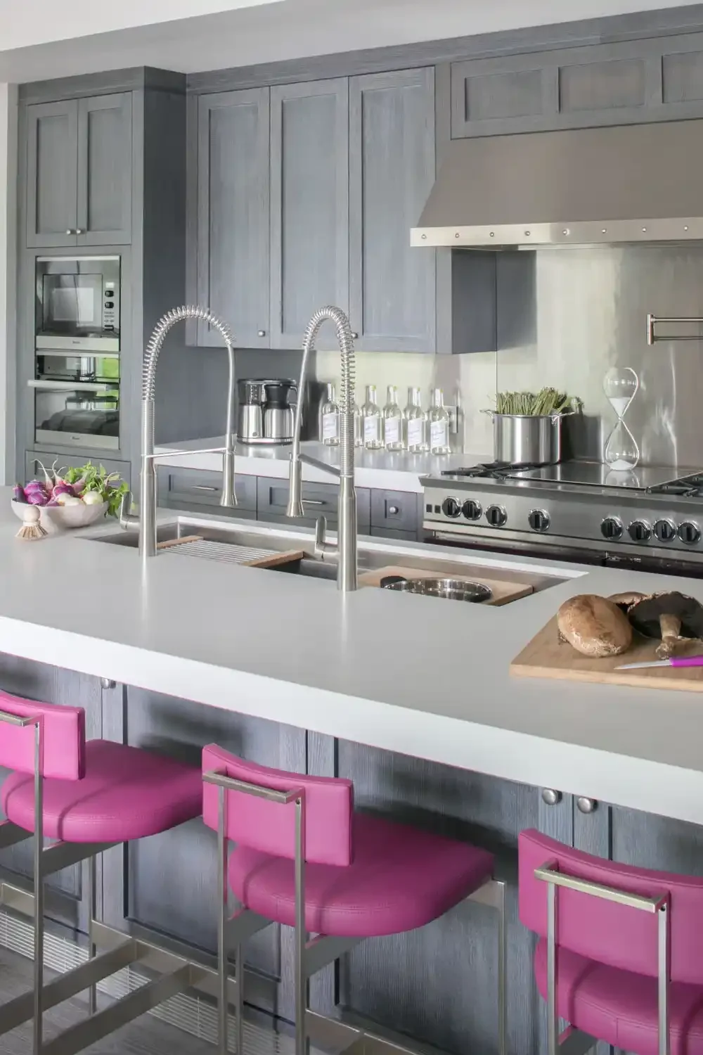 Modern kitchen with gray cabinets, stainless steel appliances, a white island counter with pink bar stools, a mounted oven, a coffee maker, bottles on the counter, and mushrooms on a cutting board.