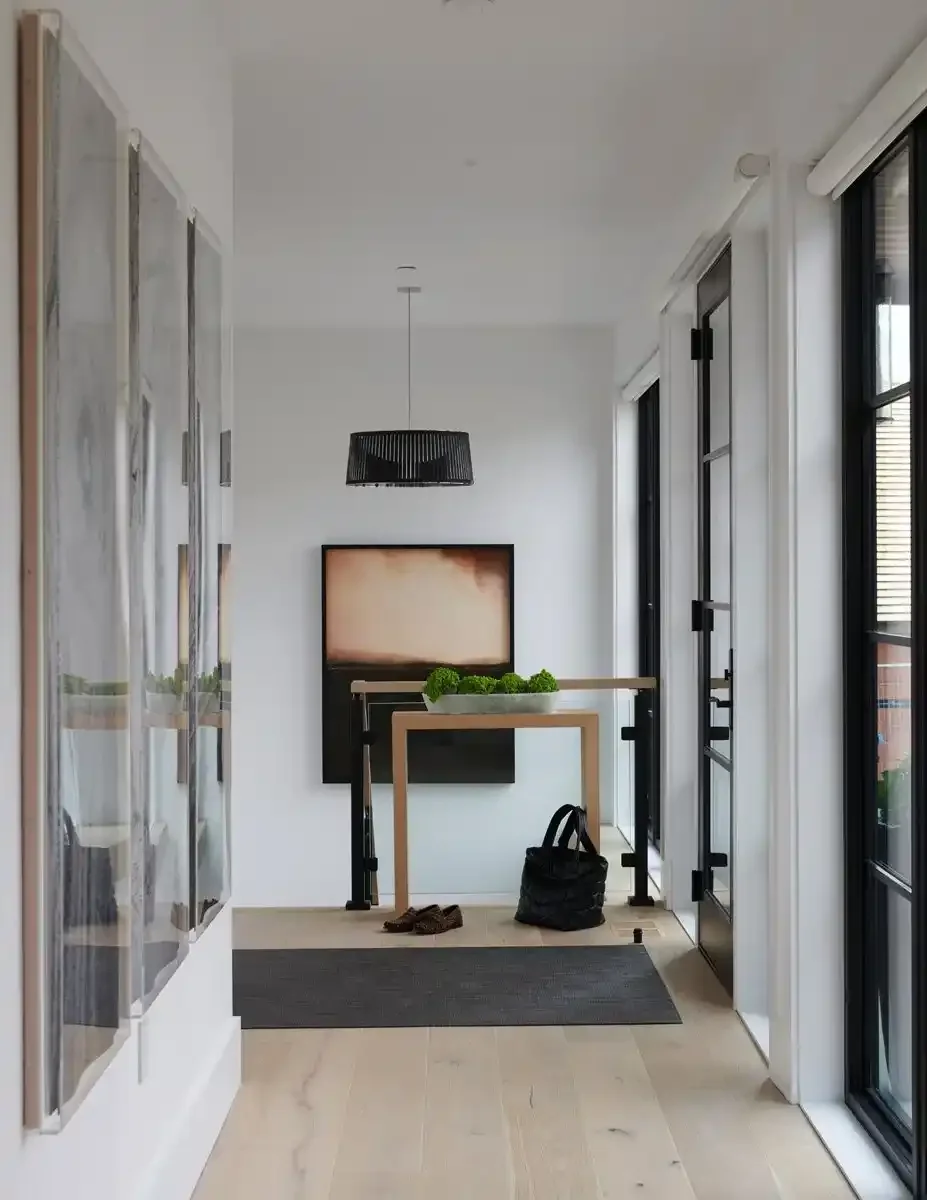Modern entryway with black-framed glass door, art piece on wall, black pendant light, black console table, decorative green moss centerpiece, black bag, brown shoes, and a gray rug on light wooden flooring.