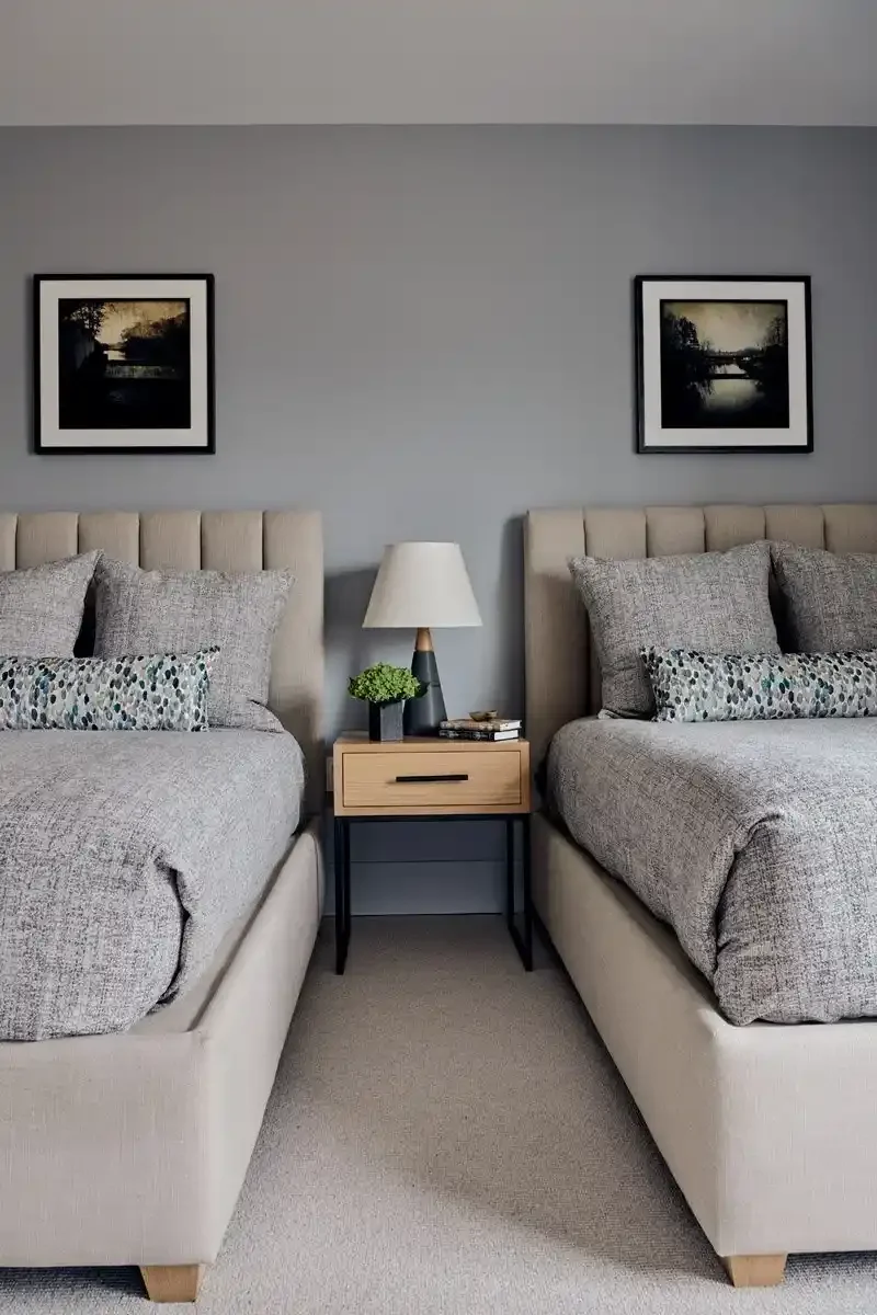 Two beige beds with gray bedding and decorative pillows, placed against a gray wall with two black-framed landscape art pictures, a wooden nightstand with a white lamp, green plant, and books between the beds.