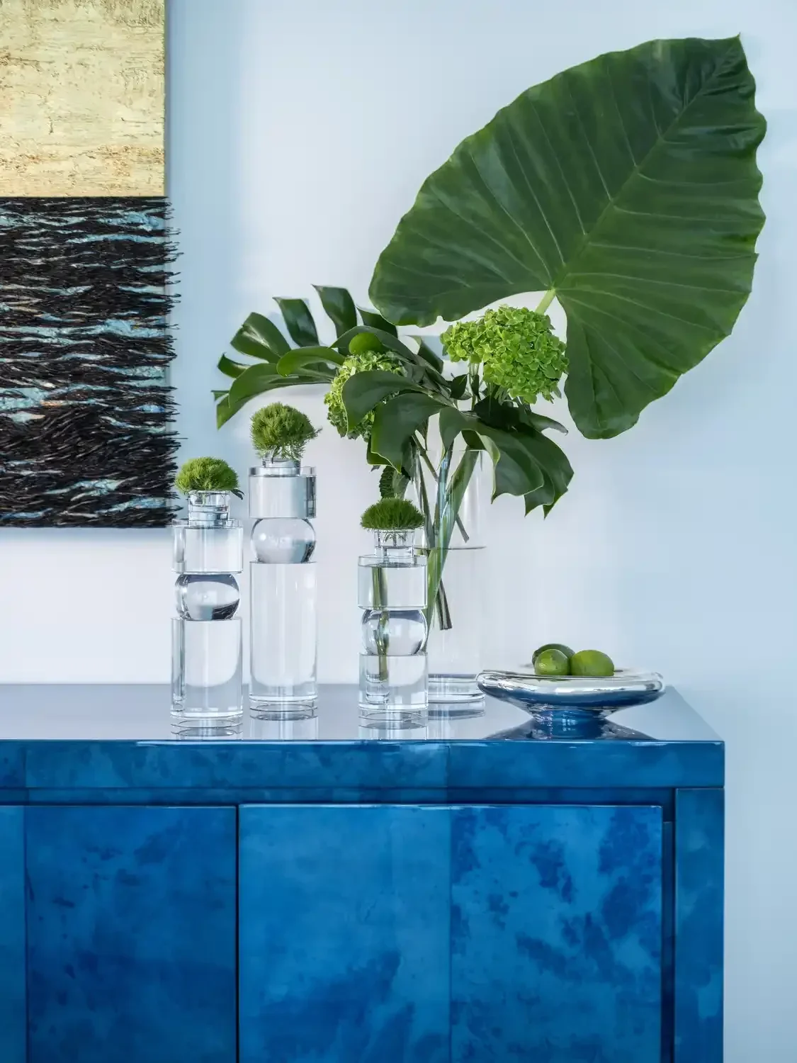 Decorative arrangement of green plants in glass vases on a blue cabinet with a dark textured artwork on the wall.