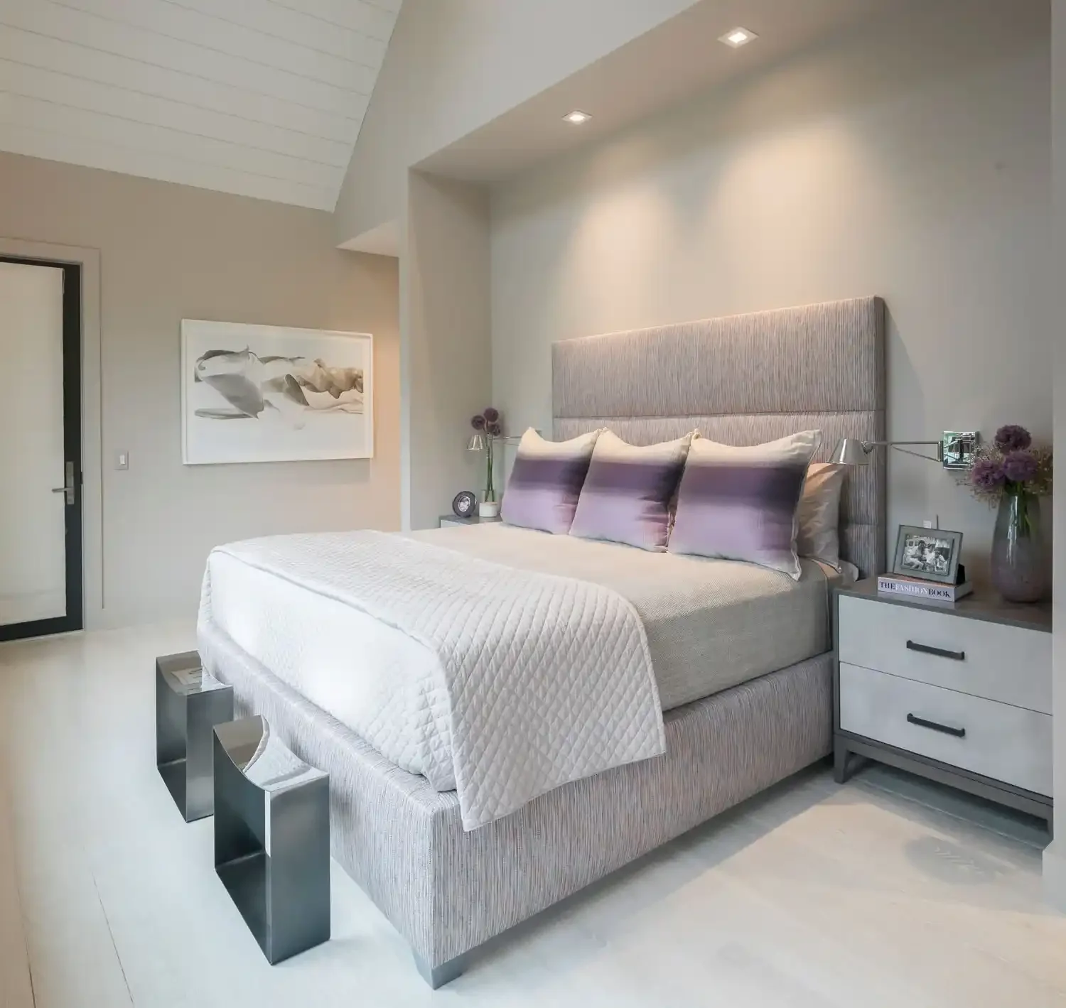 Modern bedroom with a large bed, gray upholstered headboard, and purple pillows. Nightstands on each side with lamps, flowers, and picture frames. Artwork on the wall, ceiling lights, and a white quilted bedspread.
