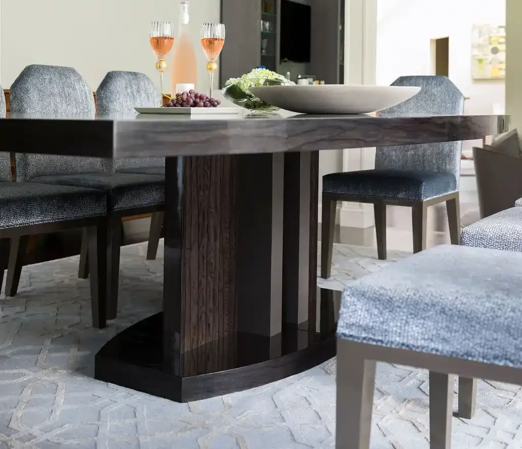 Dining room table with a modern dark wood base, surrounded by six upholstered chairs. The table has two glasses of pink champagne, a bowl, a flower arrangement, and a plate with grapes on it.
