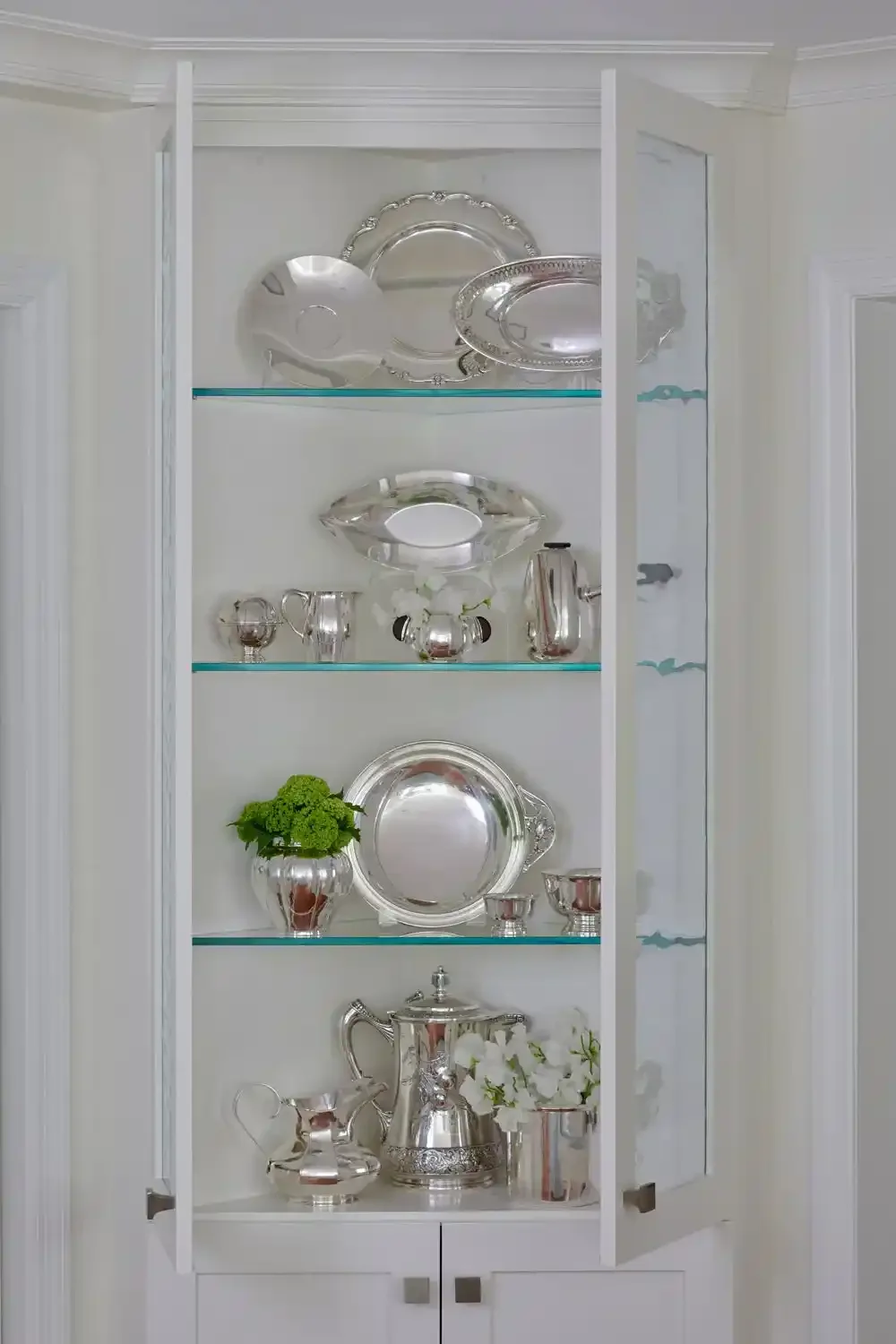 A white display cabinet with glass doors filled with silver and metallic decorative items including serving trays, pitchers, cups, vases, and a small plant with white flowers.