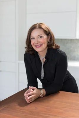 A woman with shoulder-length brown hair, wearing a black blazer, smiling and leaning on a wooden table in a bright, modern room.